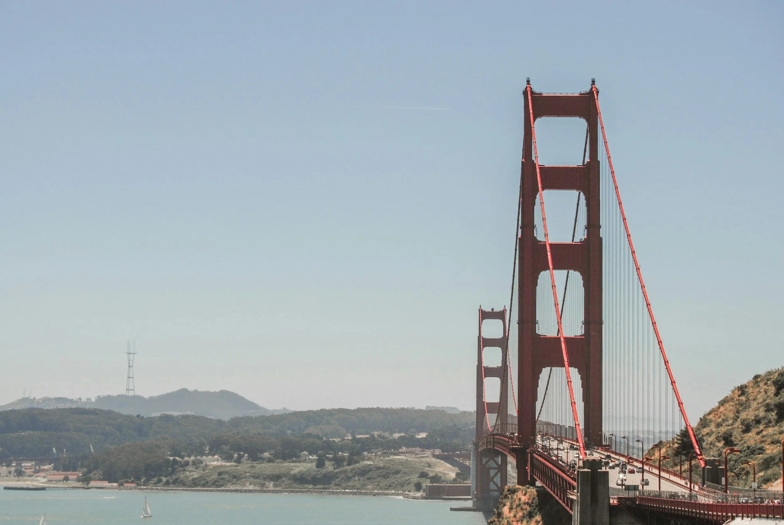 Golden Gate Bridge in San Francisco under a clear sky, with a sailboat on the water and hilly terrain in the background.