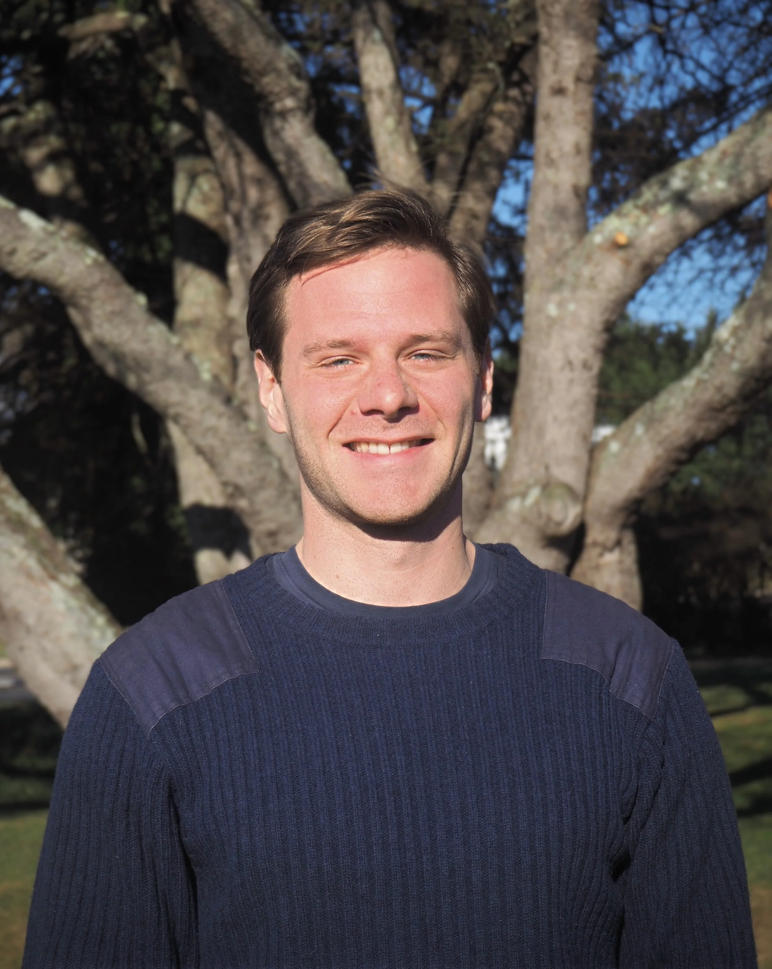 A smiling man in a dark blue sweater standing outdoors with a large, leafless tree in the background.