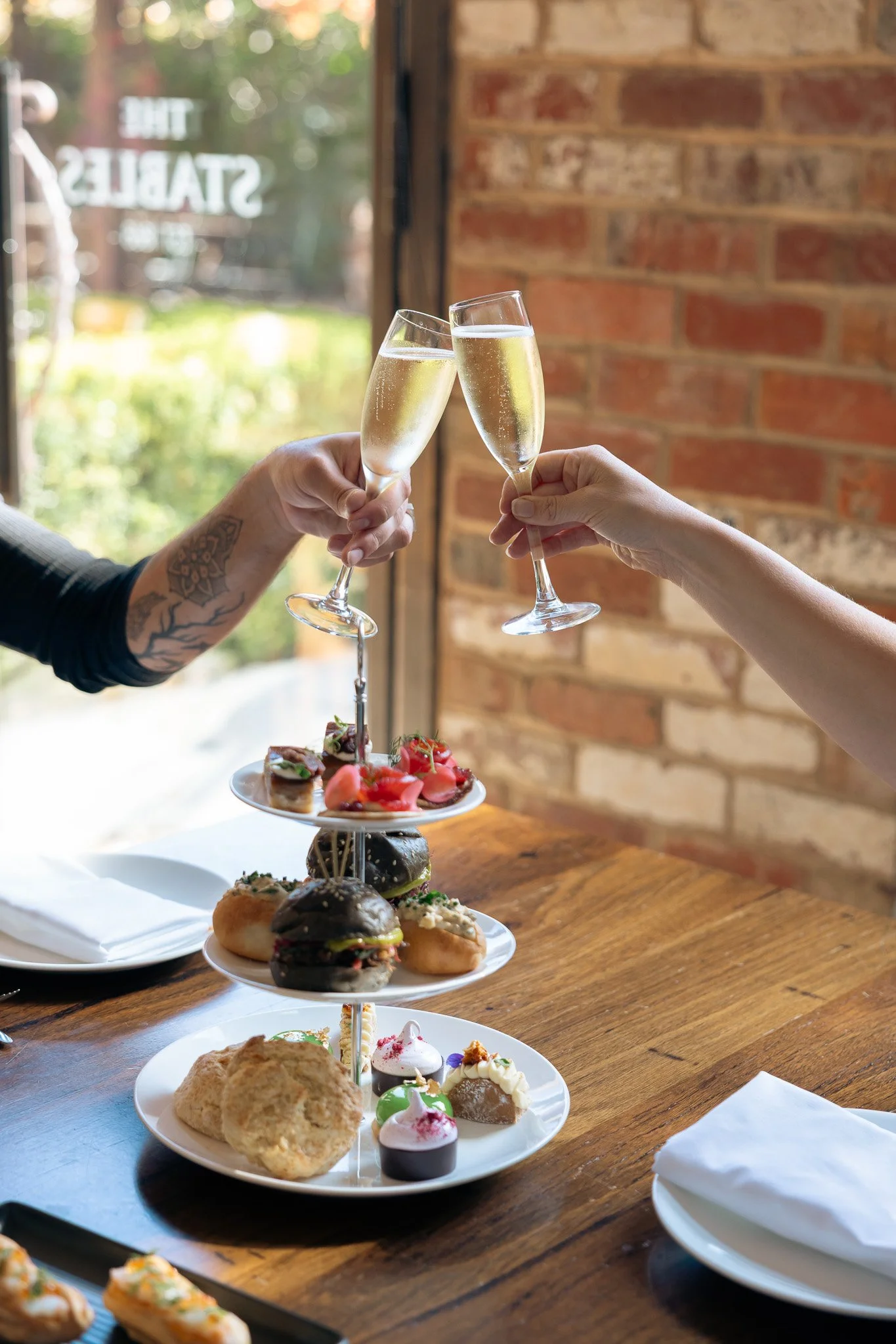 Two guests clink glasses of sparkling wine over a tiered High Tea Stand in the Stables at Stones of the Yarra Valley, featuring a selection of sweet and savoury bites.