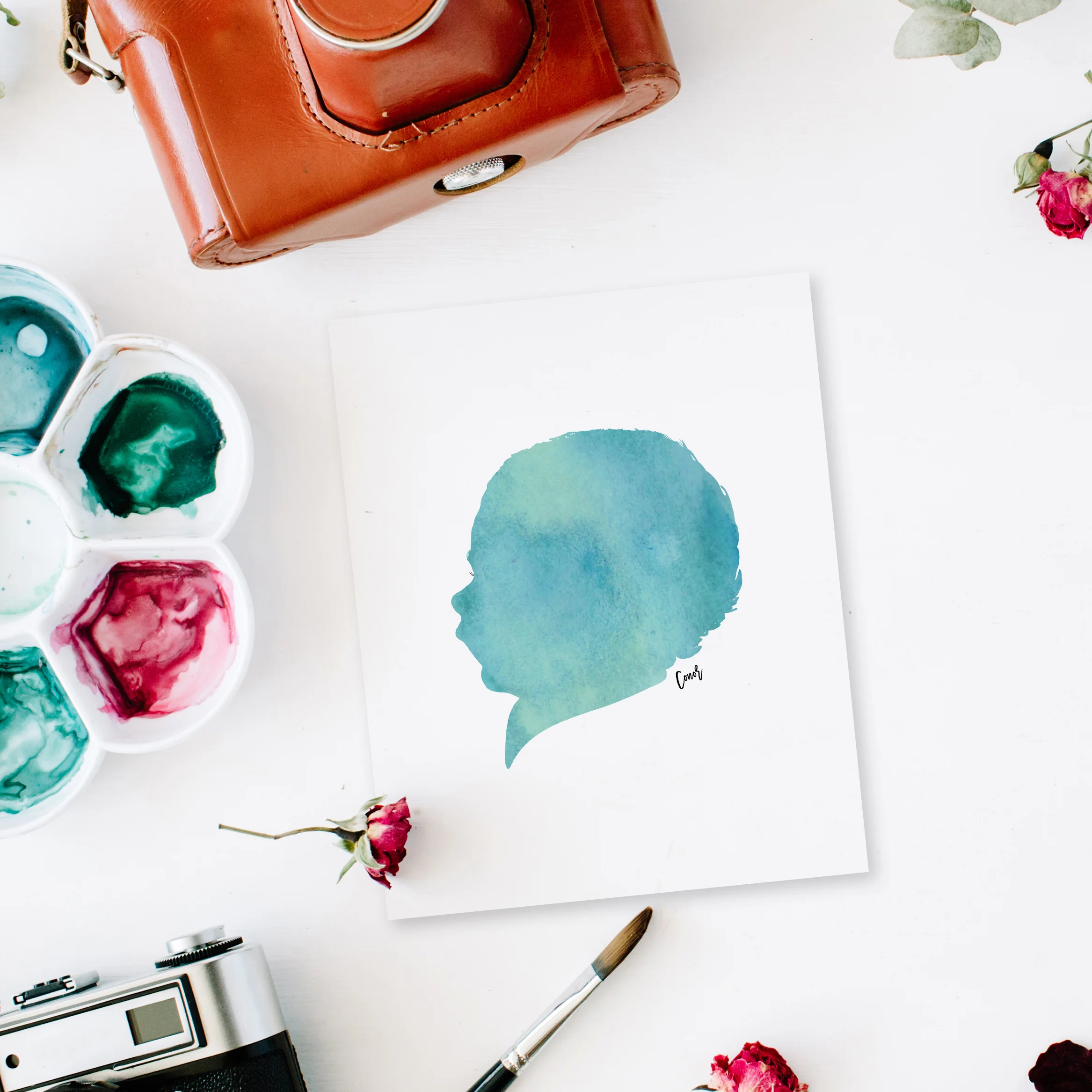 Flat lay. Artist workspace frame with vintage retro photo camera, and watercolor, red roses and eucalyptus. Top view arrangement