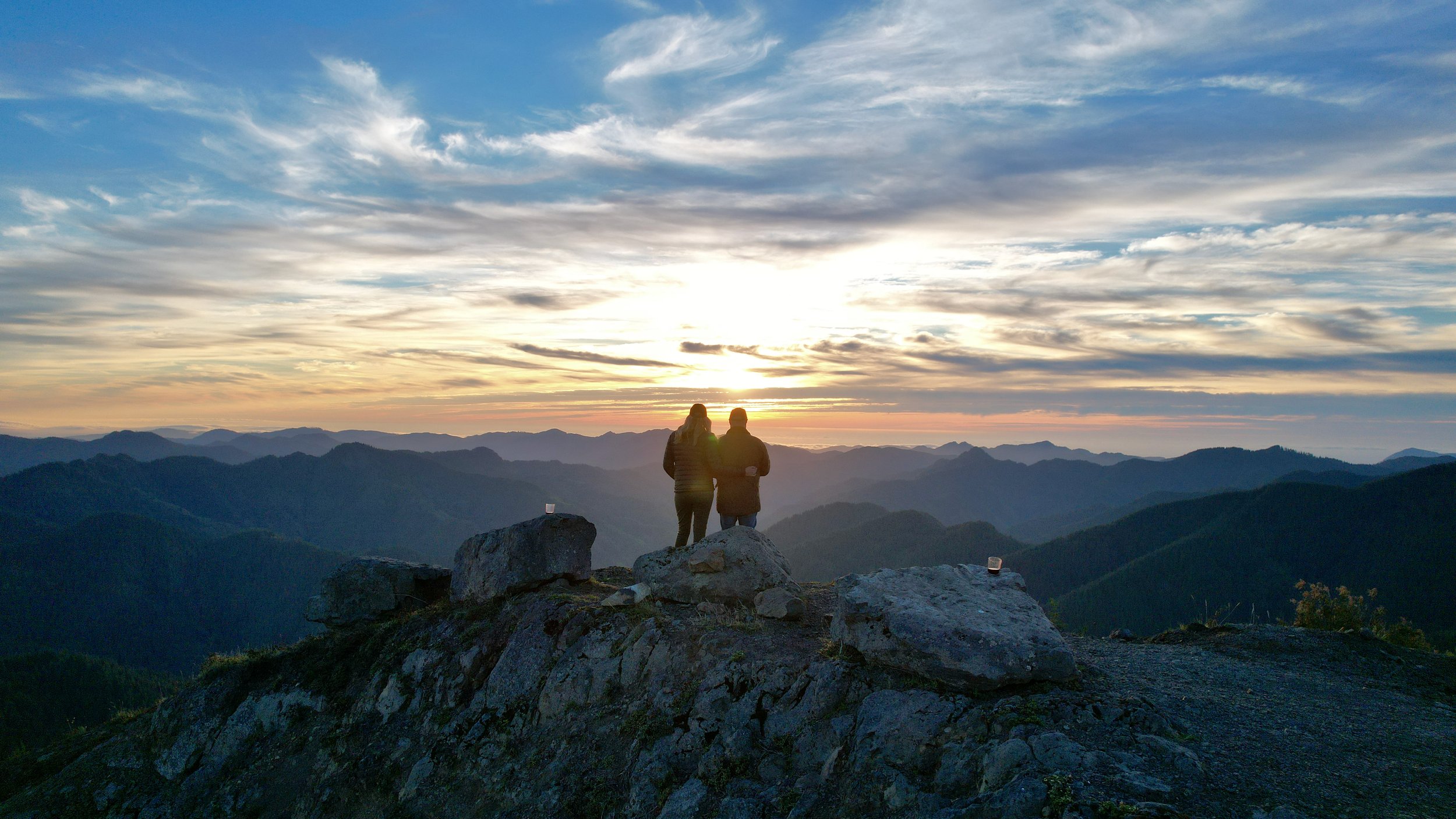 Together on a mountain summit beneath a wide sunset sky.