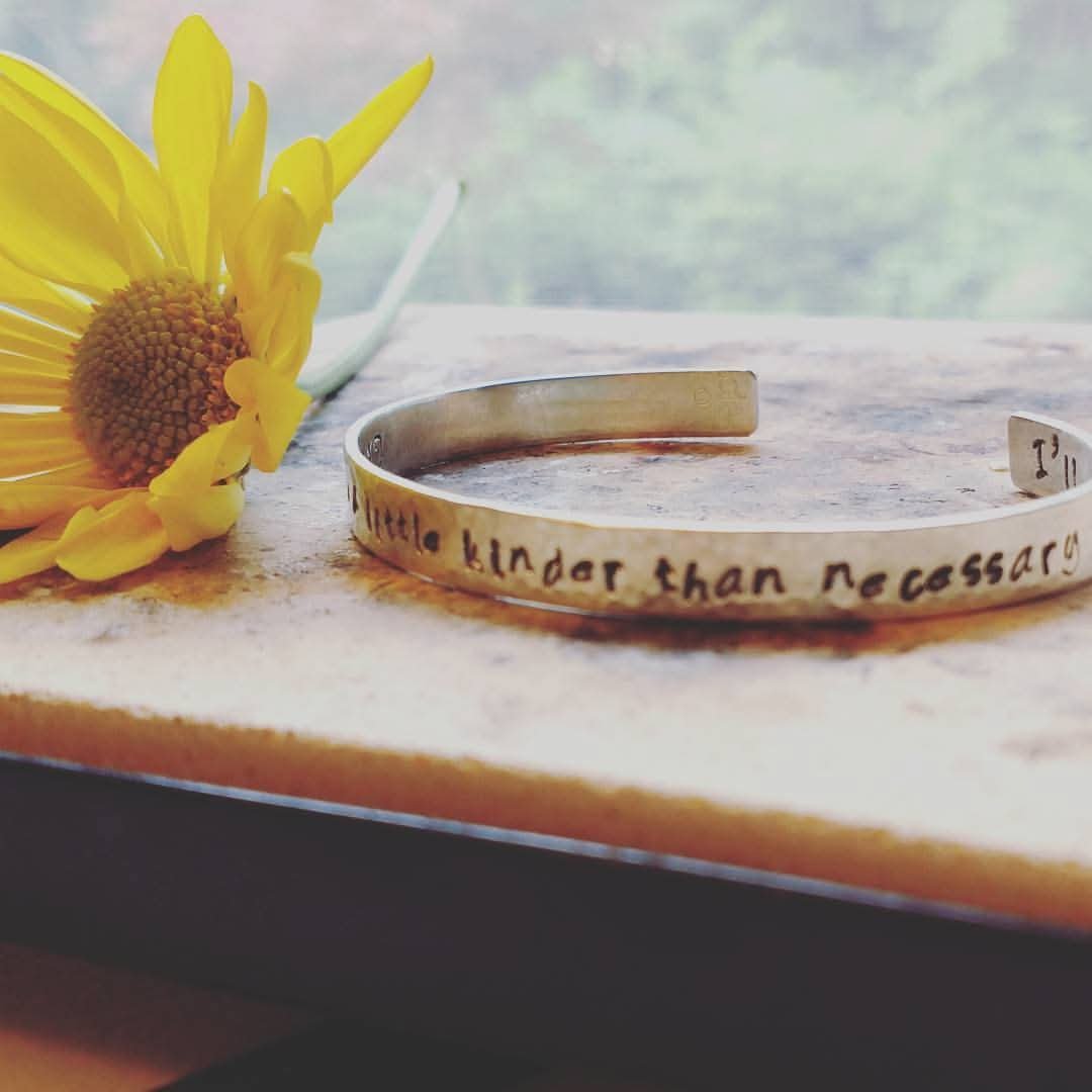 Sterling silver cuff bracelet hand stamped with “Always be a little kinder than necessary,” shown styled with a daisy on a neutral surface – handmade by MerCurios.