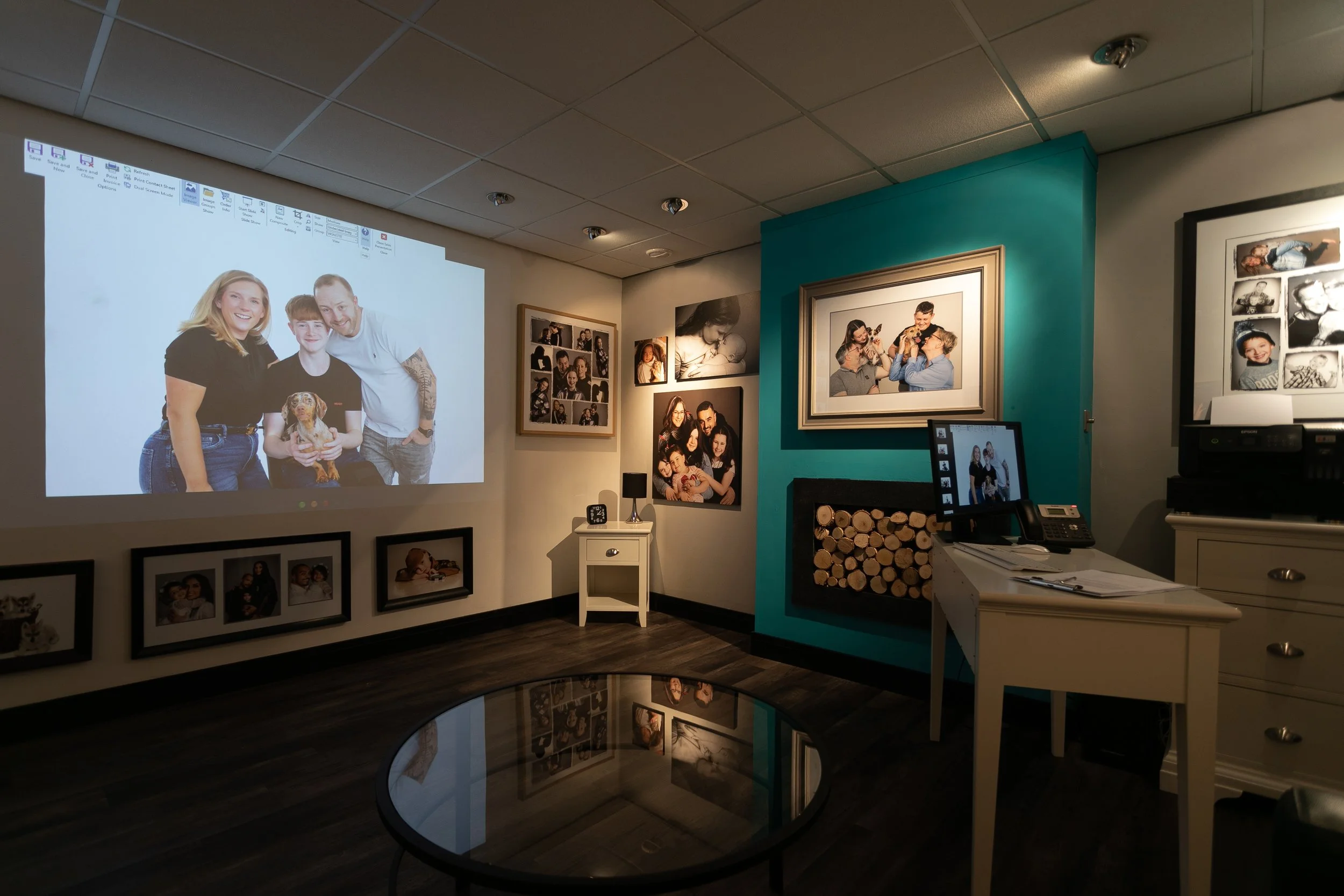 A room with a large wall projection of a family photo, small framed photos along the wall, a desk with a computer and phone, and shelves with decorative wood logs and framed pictures.
