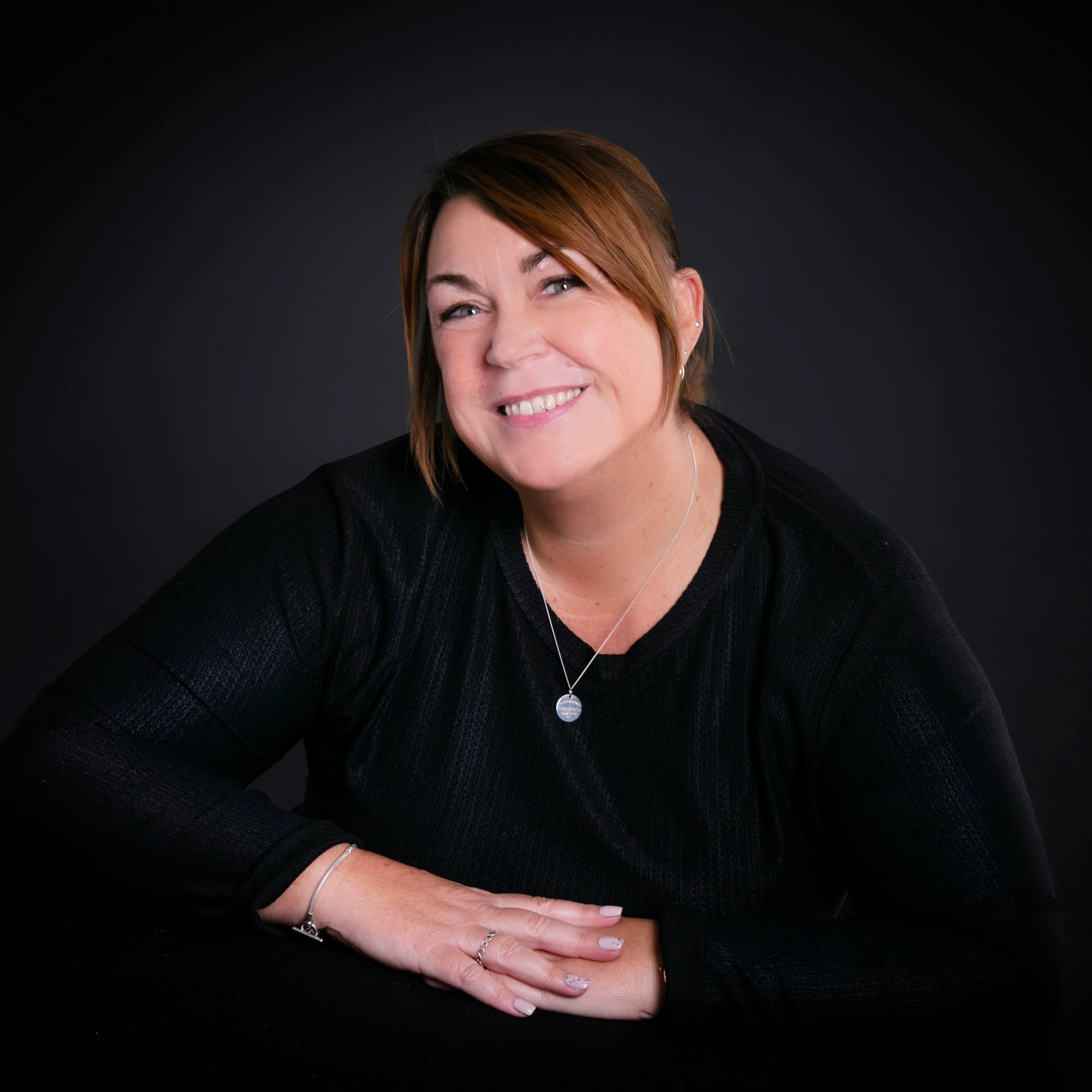 A woman with short brown hair smiling, wearing a black top, silver necklace, and rings, sitting with her arms folded on a black surface against a dark background.