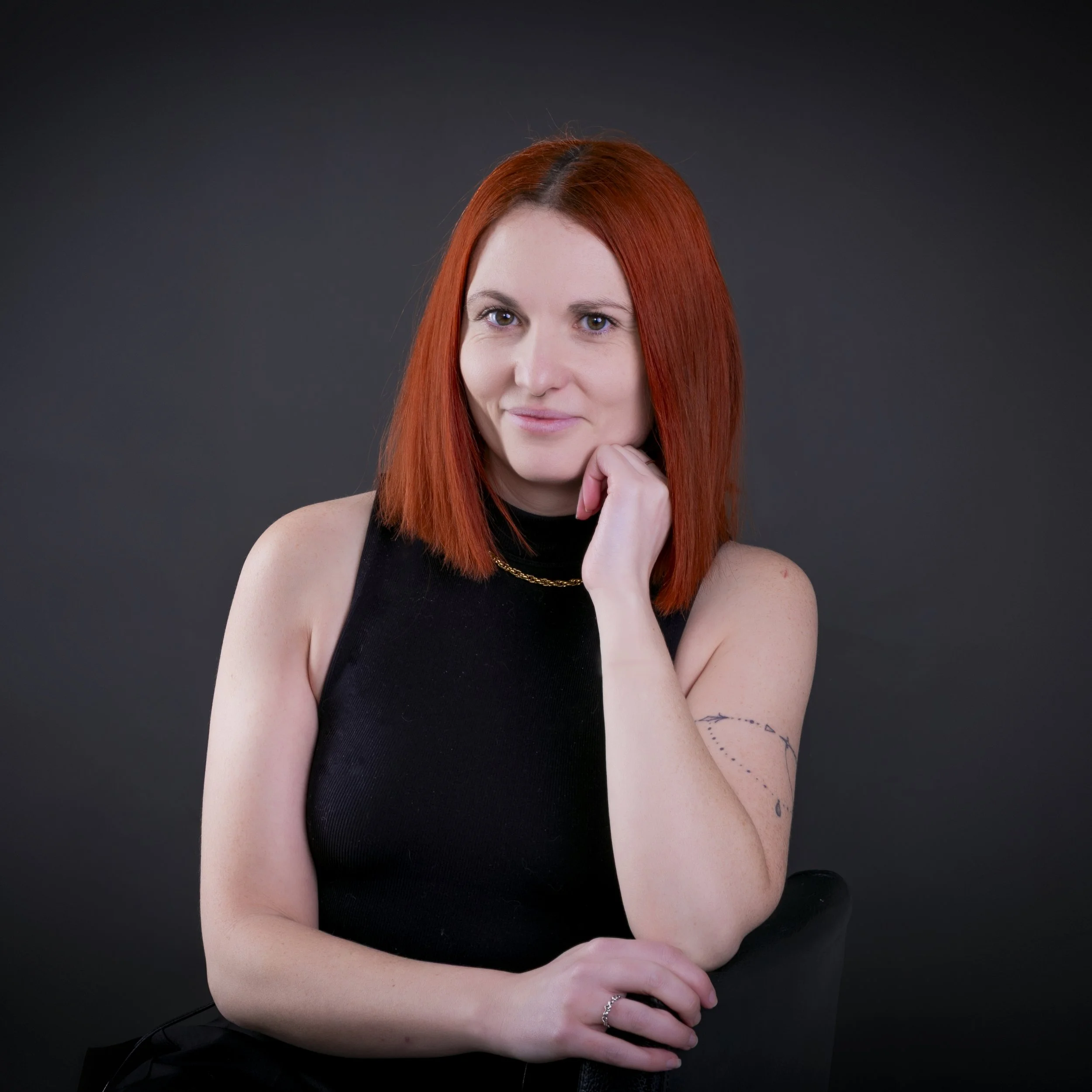 An older woman with blonde hair, glasses resting on her head, wearing a patterned gray and black top, making a playful expression with her lips pursed and eyes looking upward against a dark background.