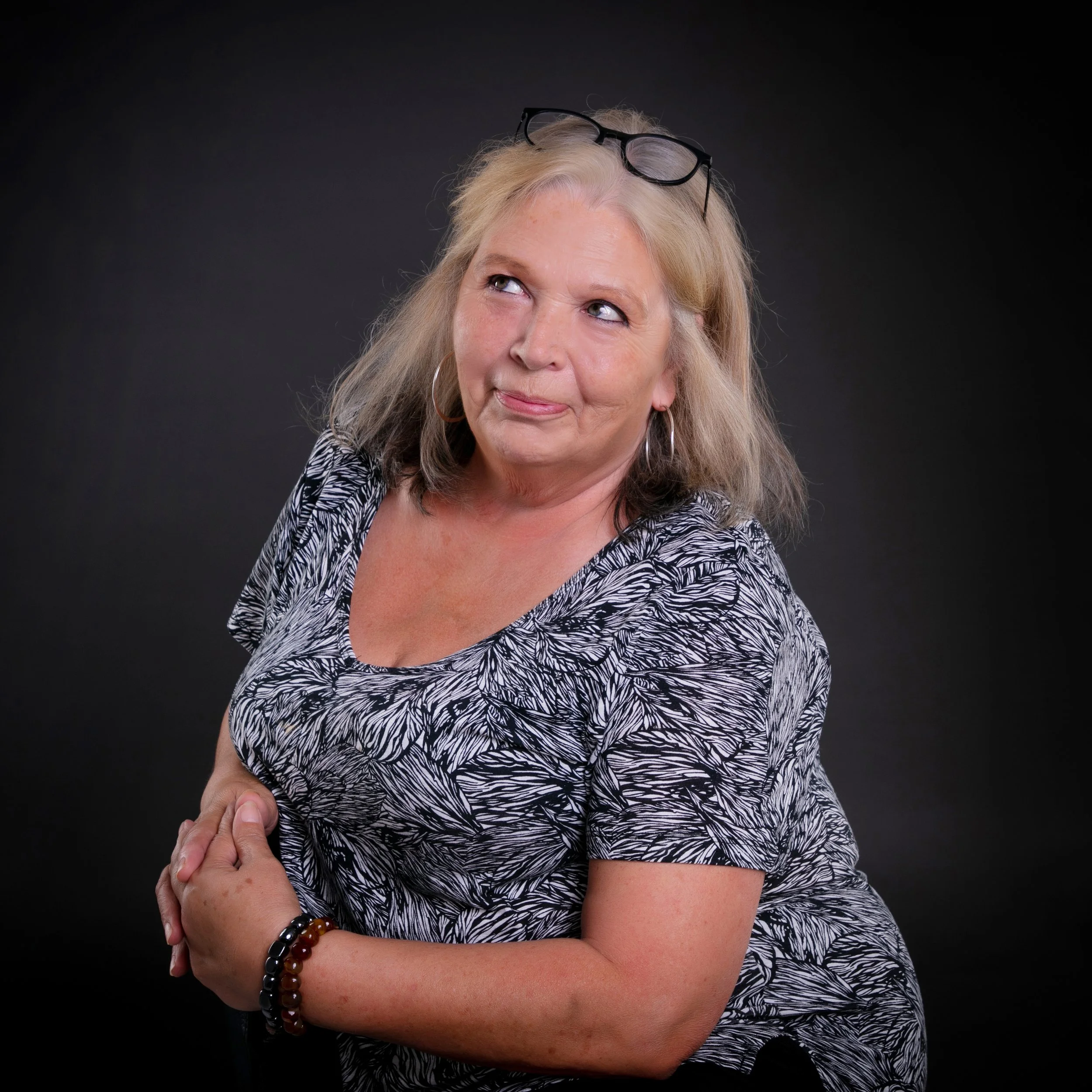 A woman sitting on a black chair against a dark background, wearing a beige sweatshirt and beige pants, with shoulder-length brown hair, looking at the camera with a slight smile.