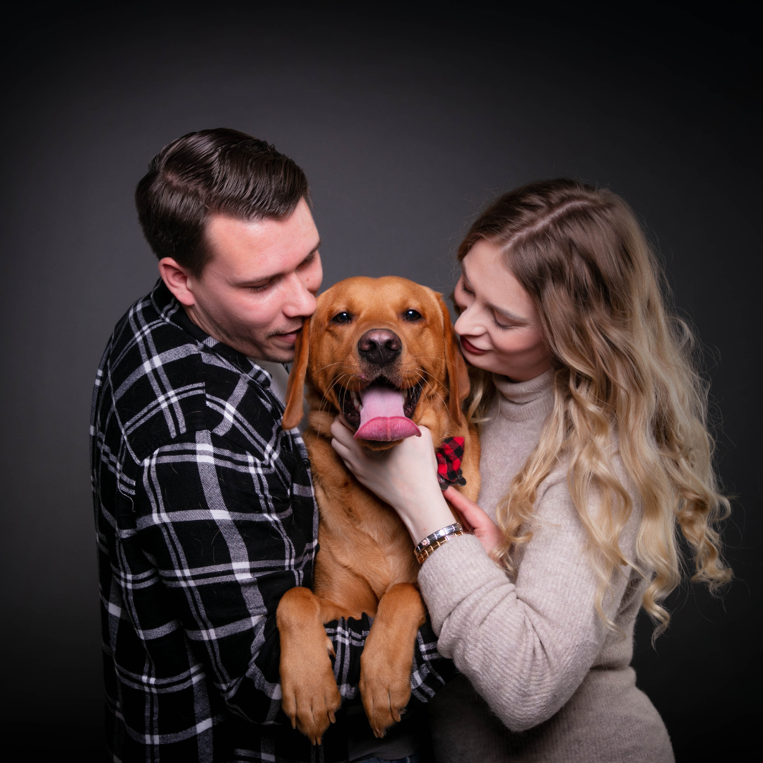 A couple holding a happy brown dog with a red and black bowtie, both leaning in towards the dog and smiling.