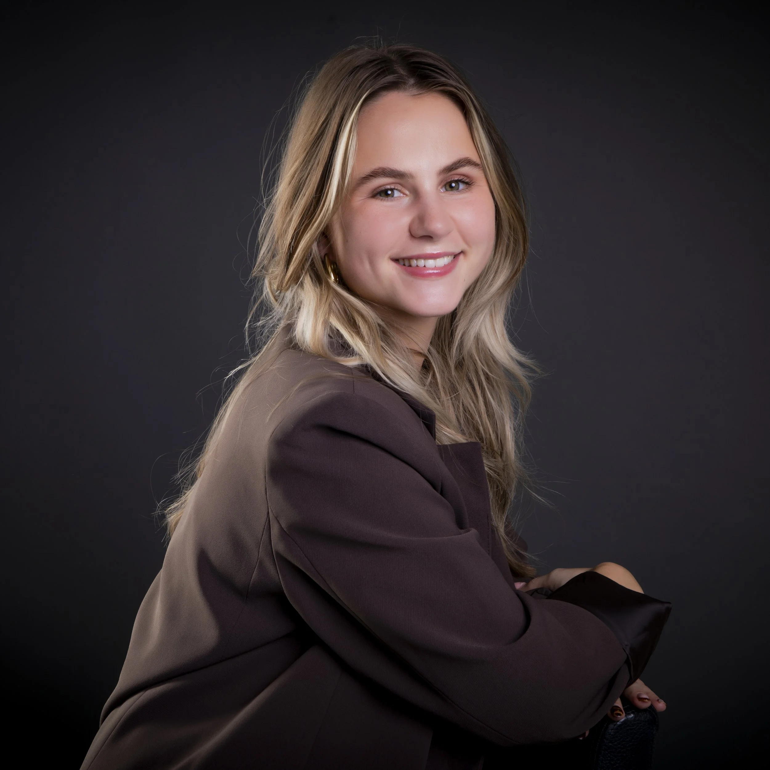 A young woman with long blonde hair smiling, wearing a teal floral dress and jewelry, sitting against a dark background.