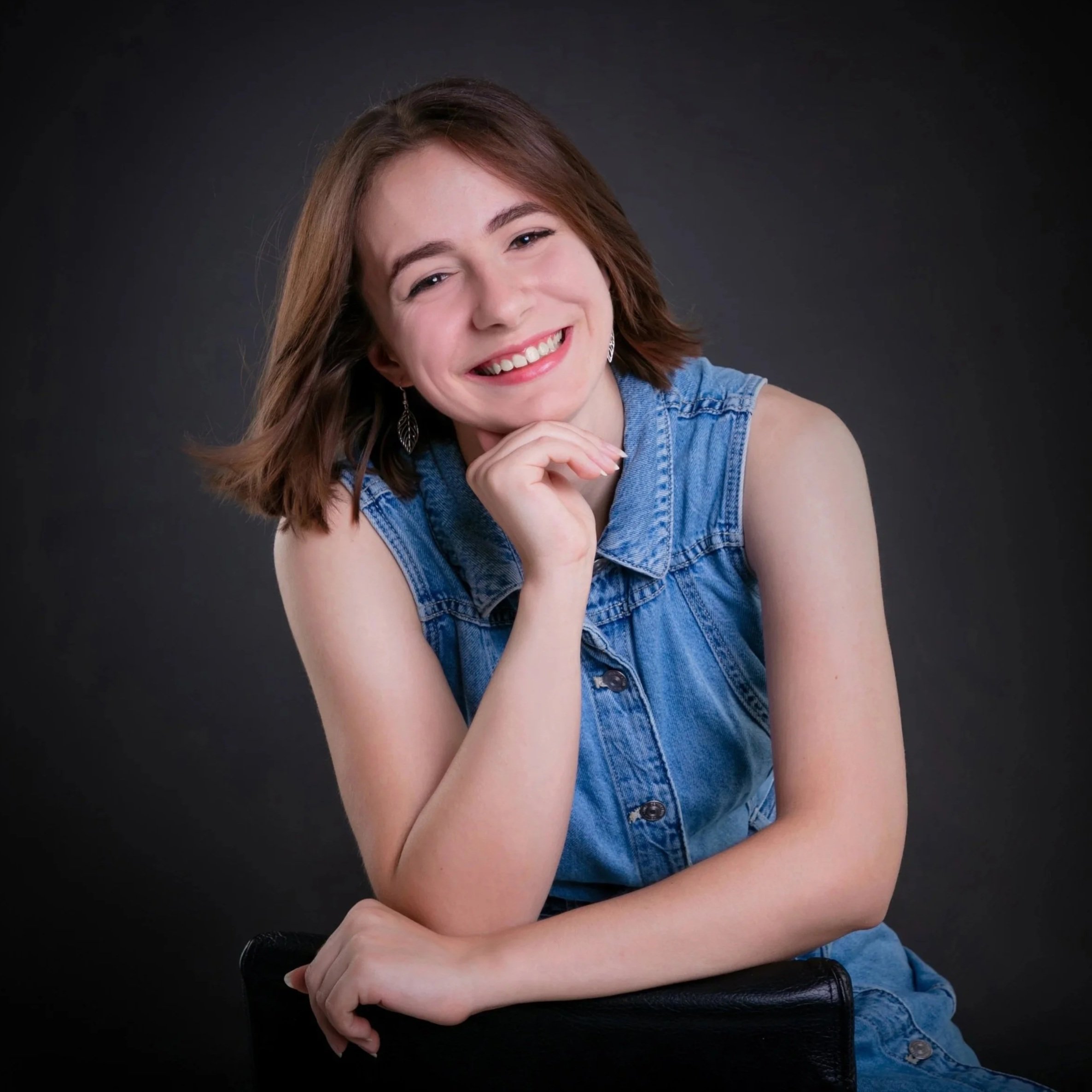 Young woman with shoulder-length brown hair smiling, wearing a sleeveless denim shirt, sitting against a black background.