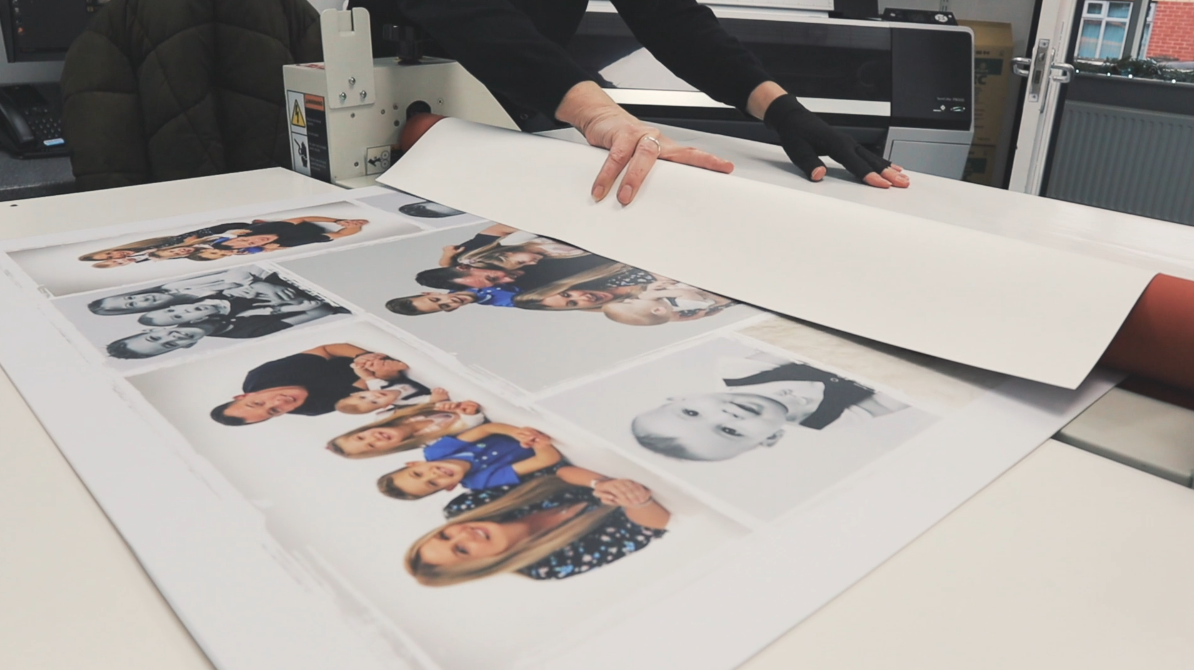 Hands unrolling a large sheet of paper with printed photographs of people on a table in a print shop or photo lab.