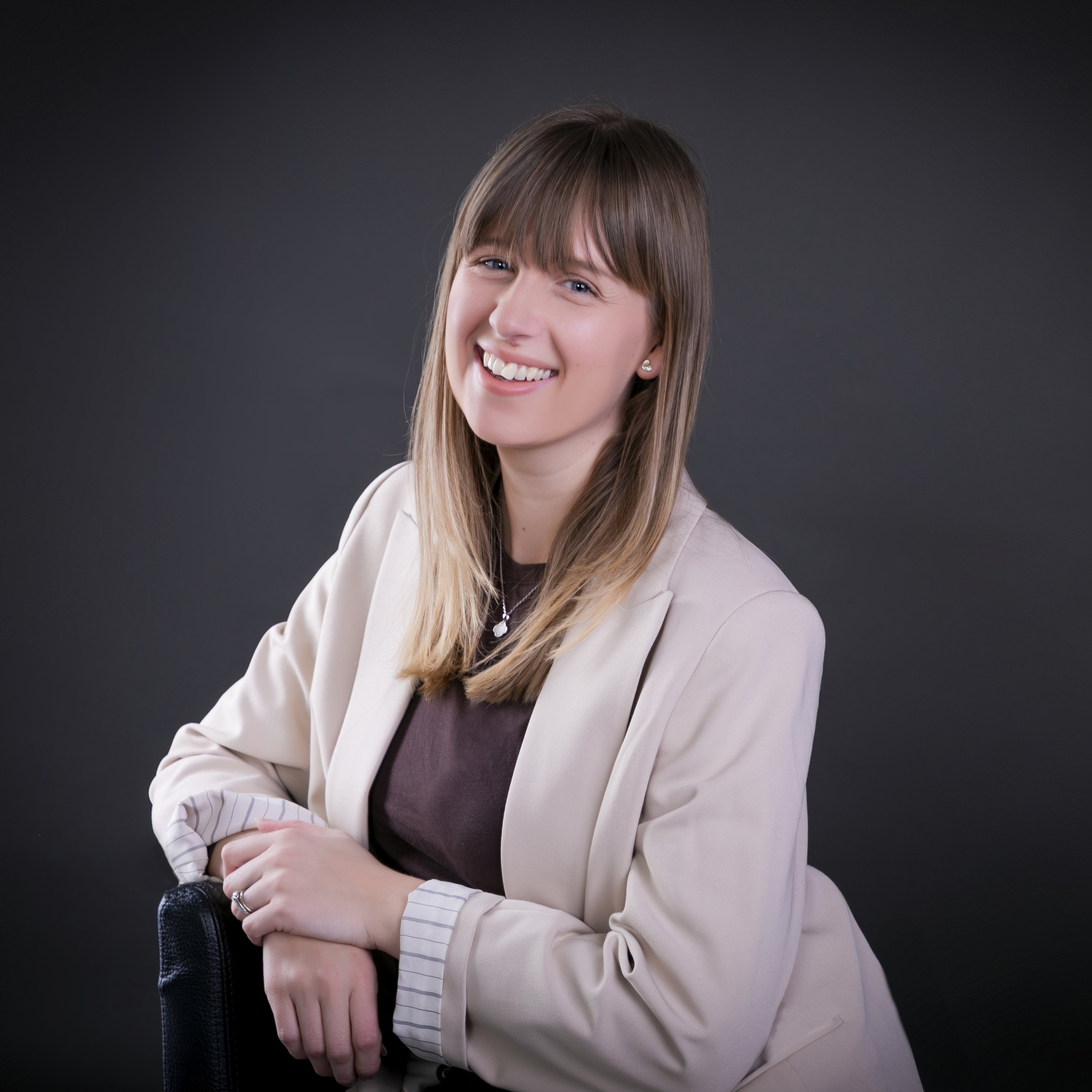 Smiling woman with long light brown hair wearing a light-colored blouse against a dark background.