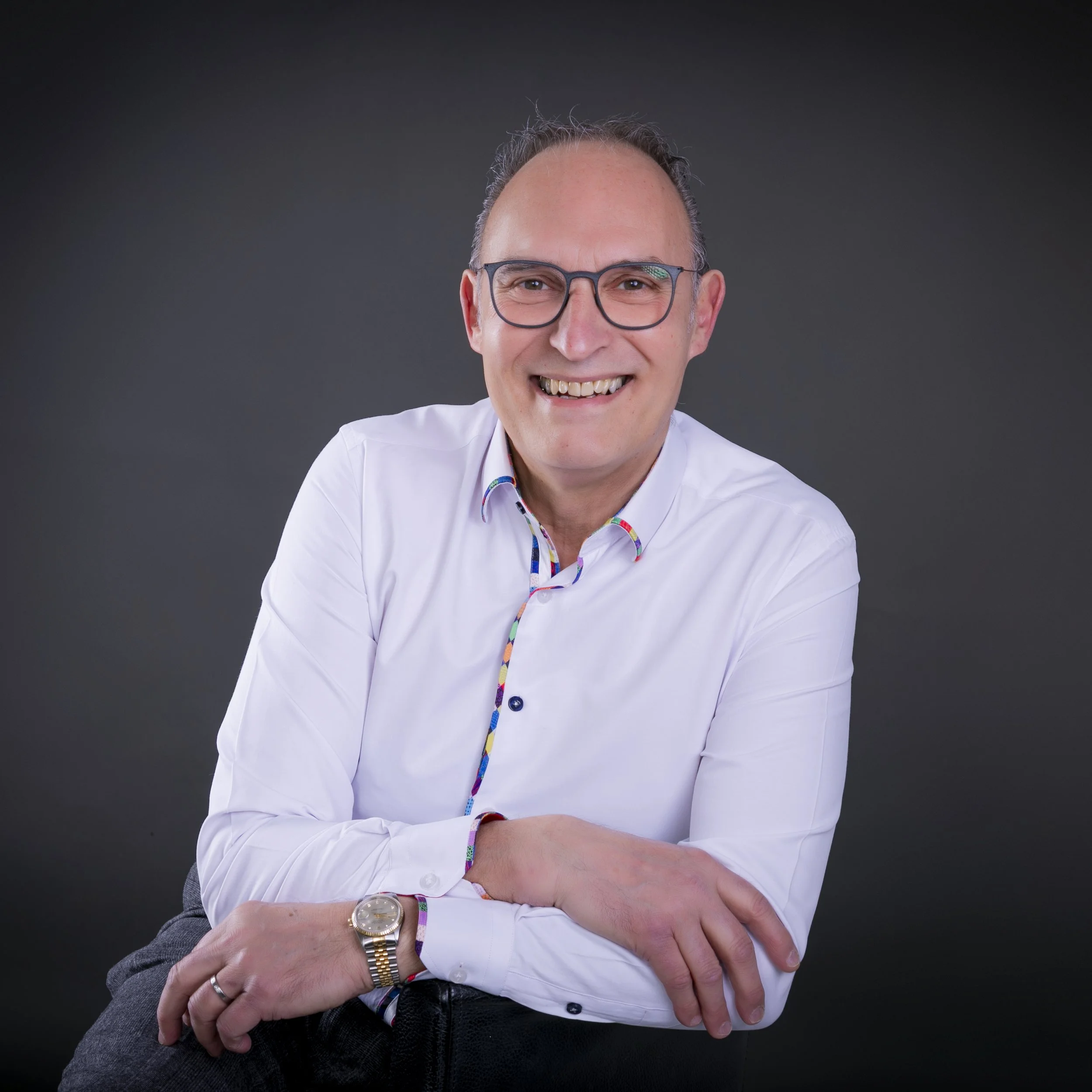 Headshot of a middle-aged man wearing glasses, a gray suit, white shirt, and a colorful tie, smiling against a dark background.