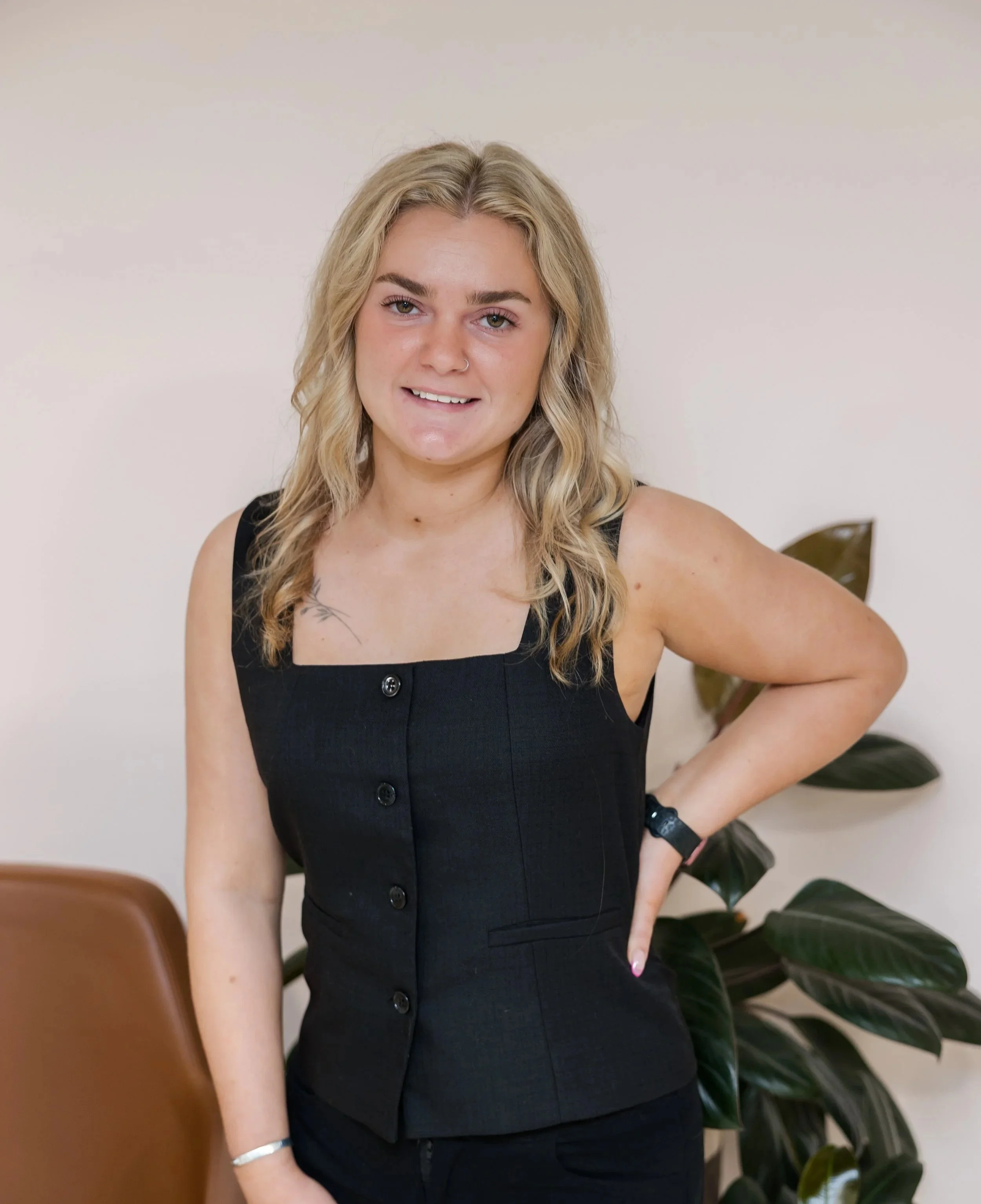 A young woman with blonde hair wearing a black sleeveless top and wristwatch, standing indoors next to a potted plant and a brown chair.