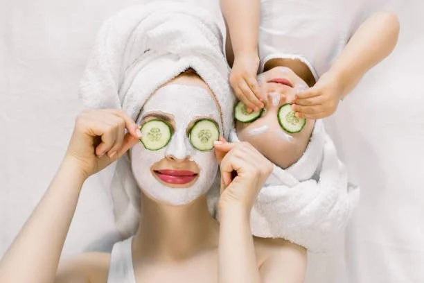 A woman with a face mask and towel on her head holds cucumber slices over her eyes, while a child beside her places cucumber slices over their eyes and forehead.