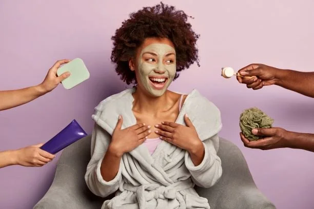 Woman with a facial mask, smiling, sitting in a chair, surrounded by hands holding beauty and skincare products and tools.