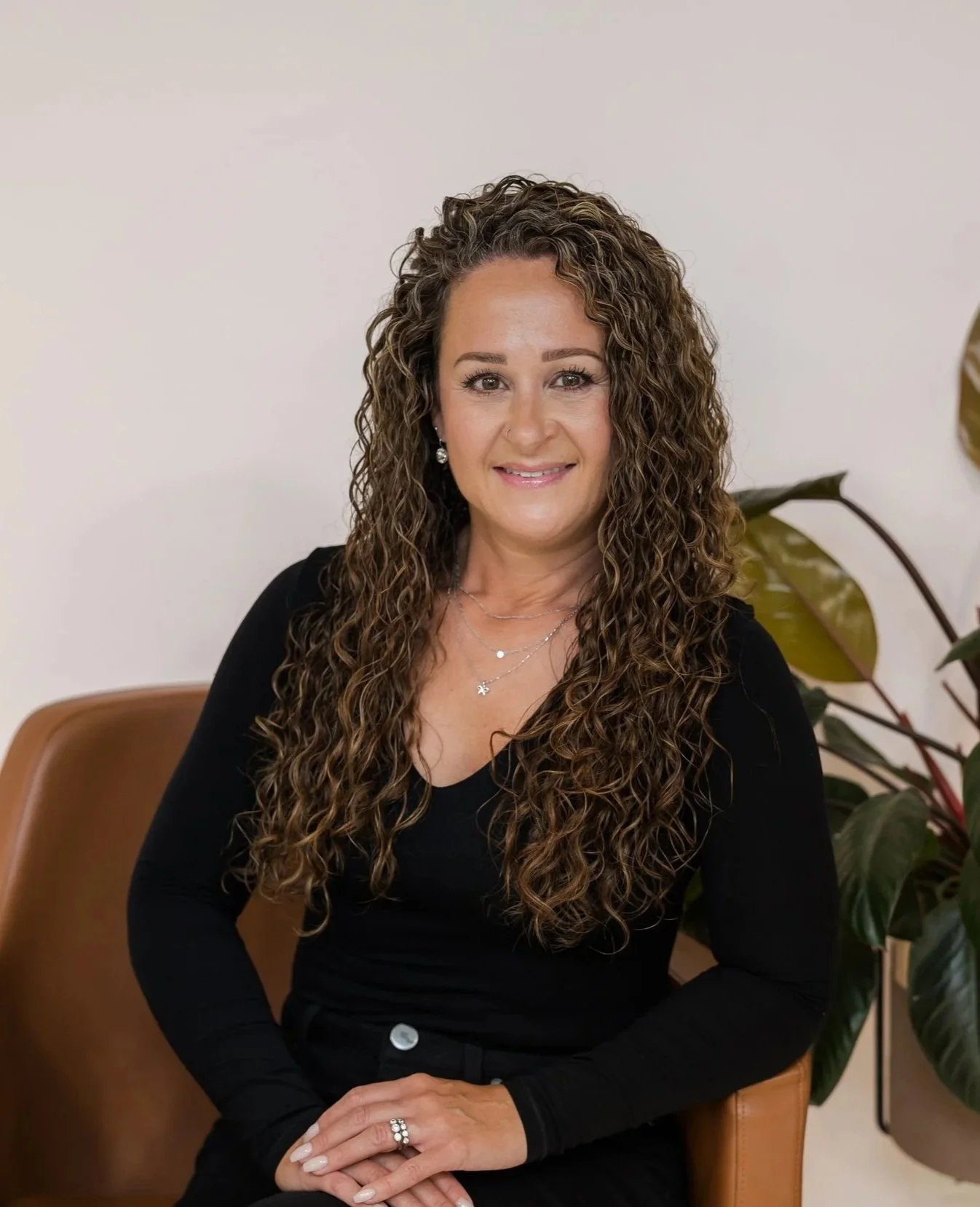 A woman with curly brown hair, wearing a black long-sleeve shirt, jewelry, and sitting on a tan chair, smiling at the camera, with a plant in the background.