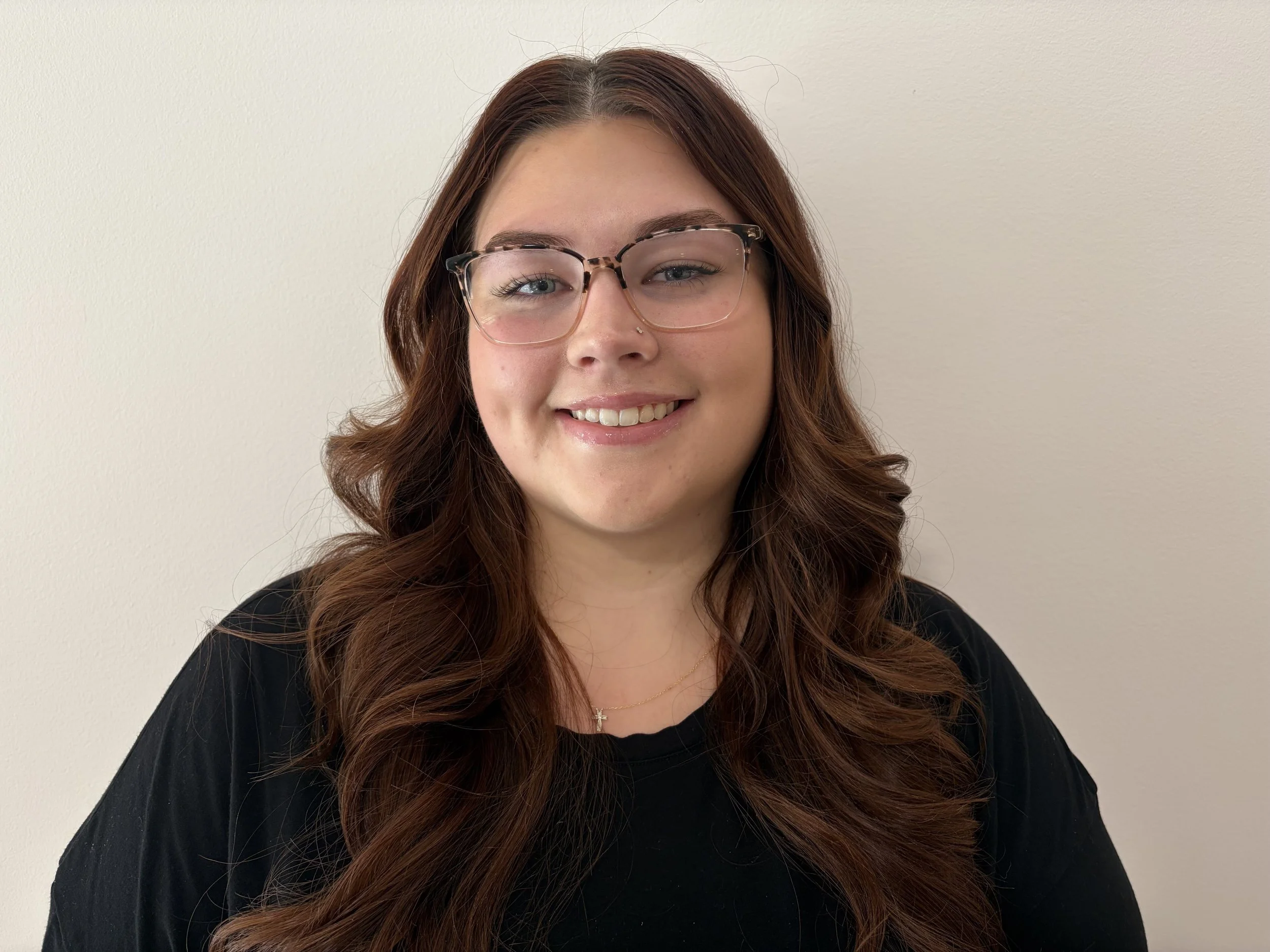 Young woman with wavy brown hair, glasses, and a black shirt, smiling against a plain light-colored wall.