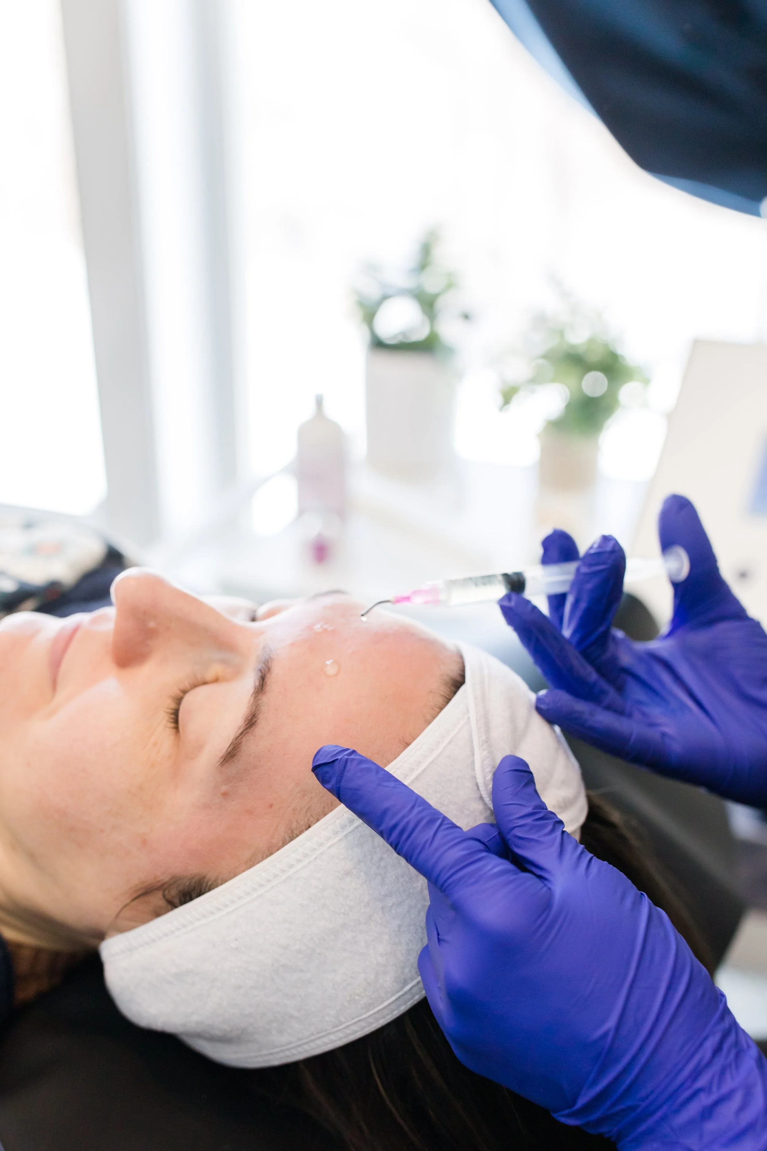 A person receiving a cosmetic injection on her forehead while lying down with a white headband, with gloved hands administering the treatment, in a bright room with plants and medical equipment in the background.