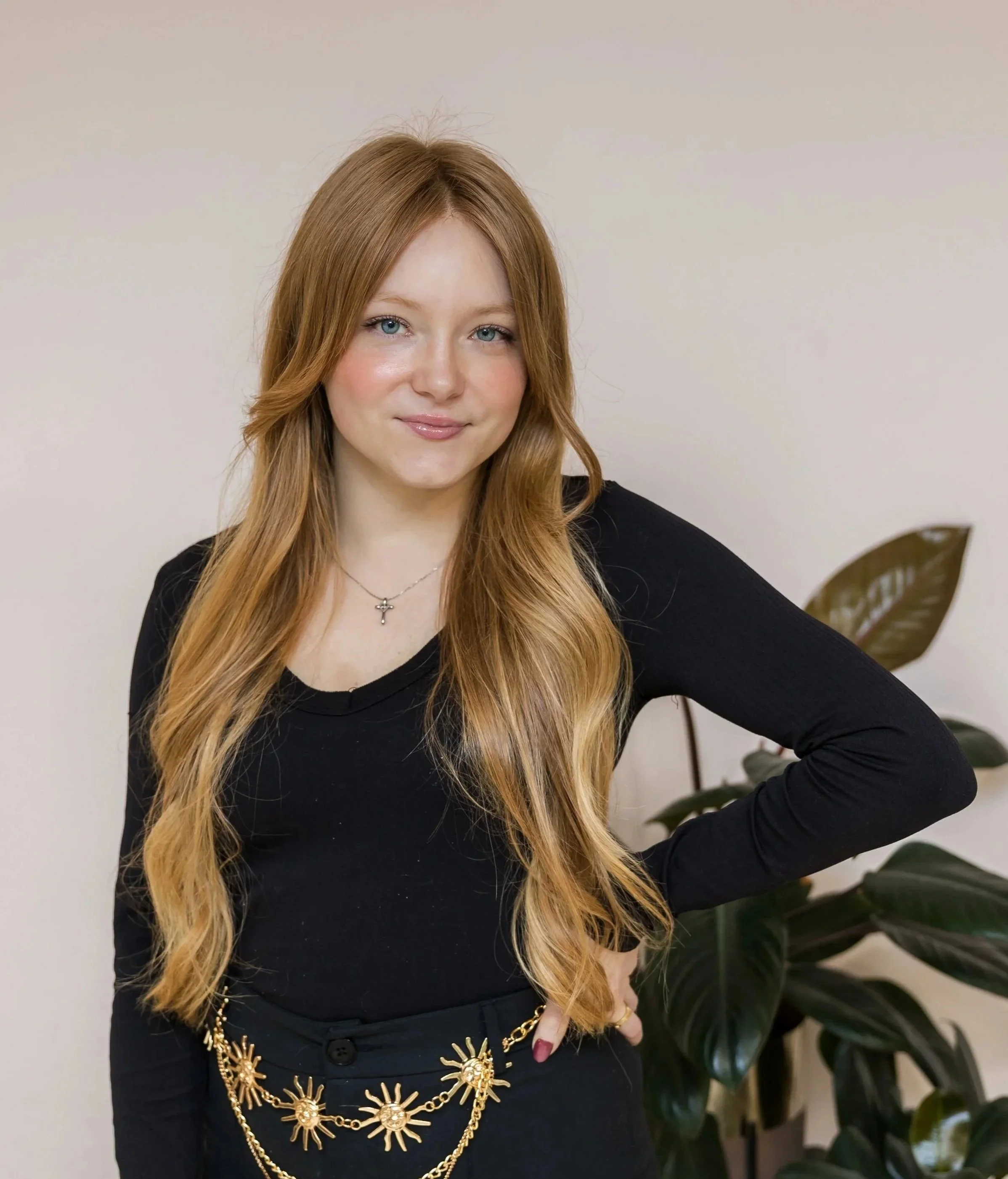 A young woman with long red hair and blue eyes, wearing a black top and a sun-themed gold necklace, standing in front of a plain light-colored wall with a large green plant in the background.