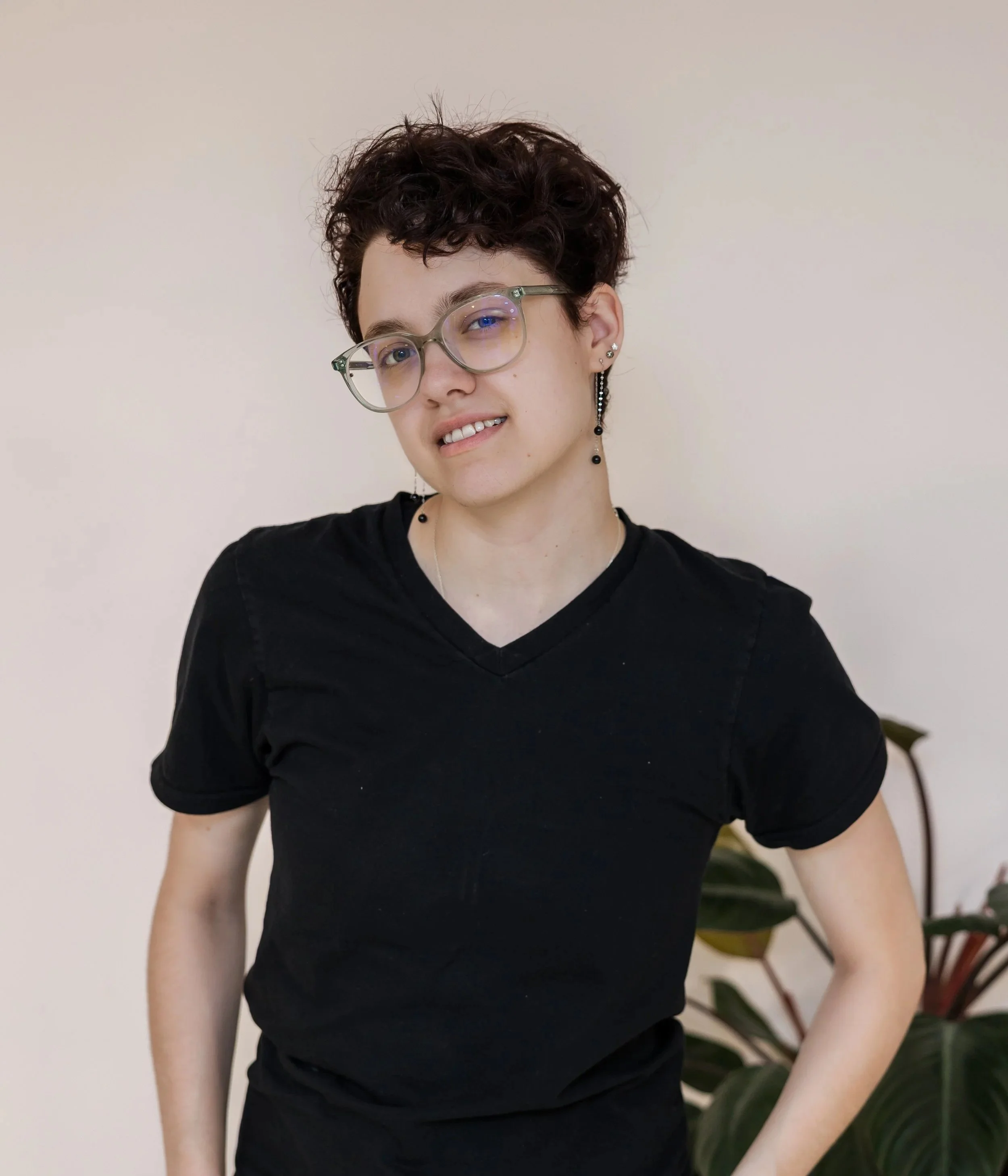 A person with short, curly dark hair and light skin wearing glasses and multiple earrings, smiling while posing in front of a bare wall with some green leaves visible on the right side.
