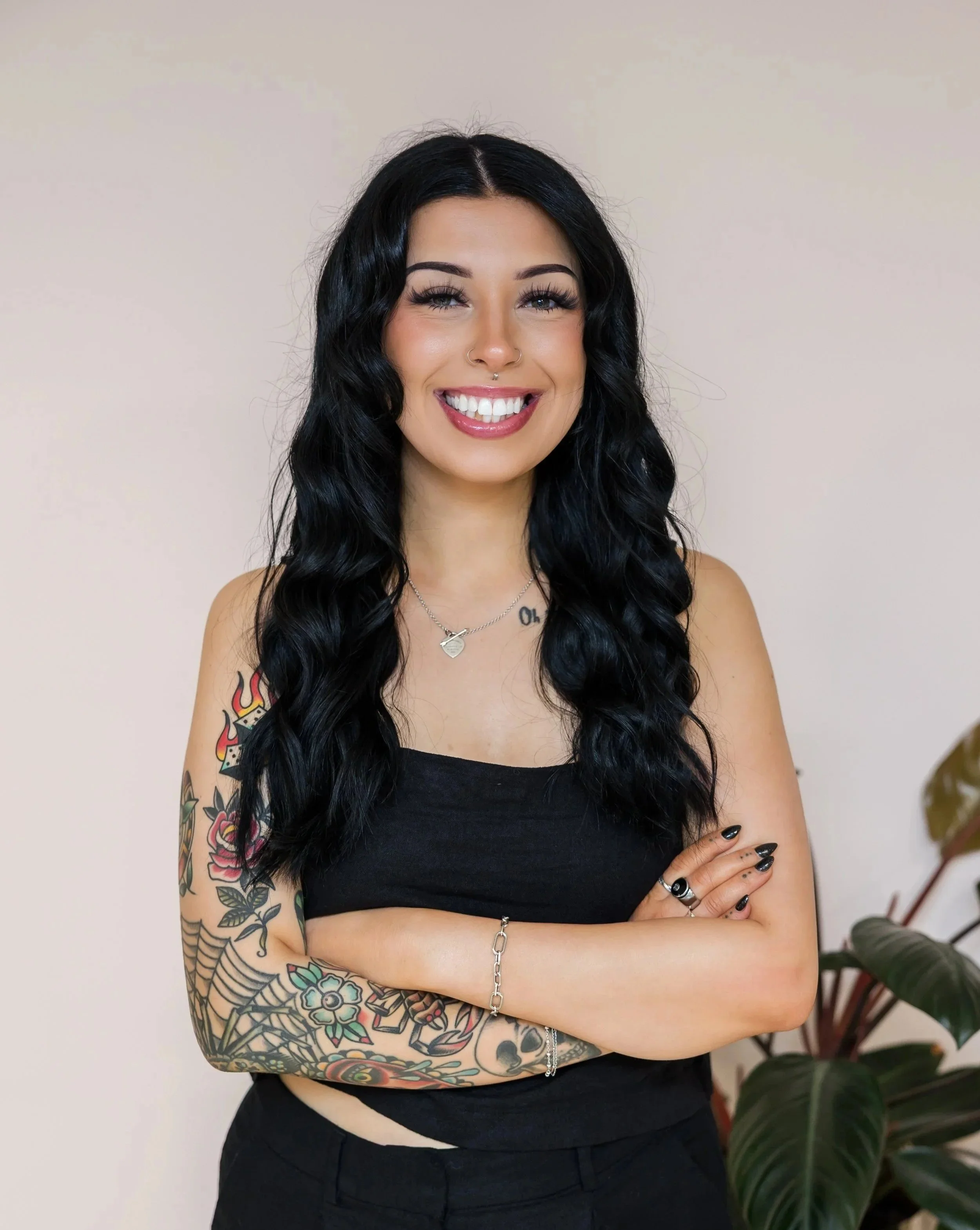 A woman with long, wavy black hair, tattoos on her arms, smiling with arms crossed, wearing a black sleeveless top, and standing in front of a light-colored wall with a plant to her right.