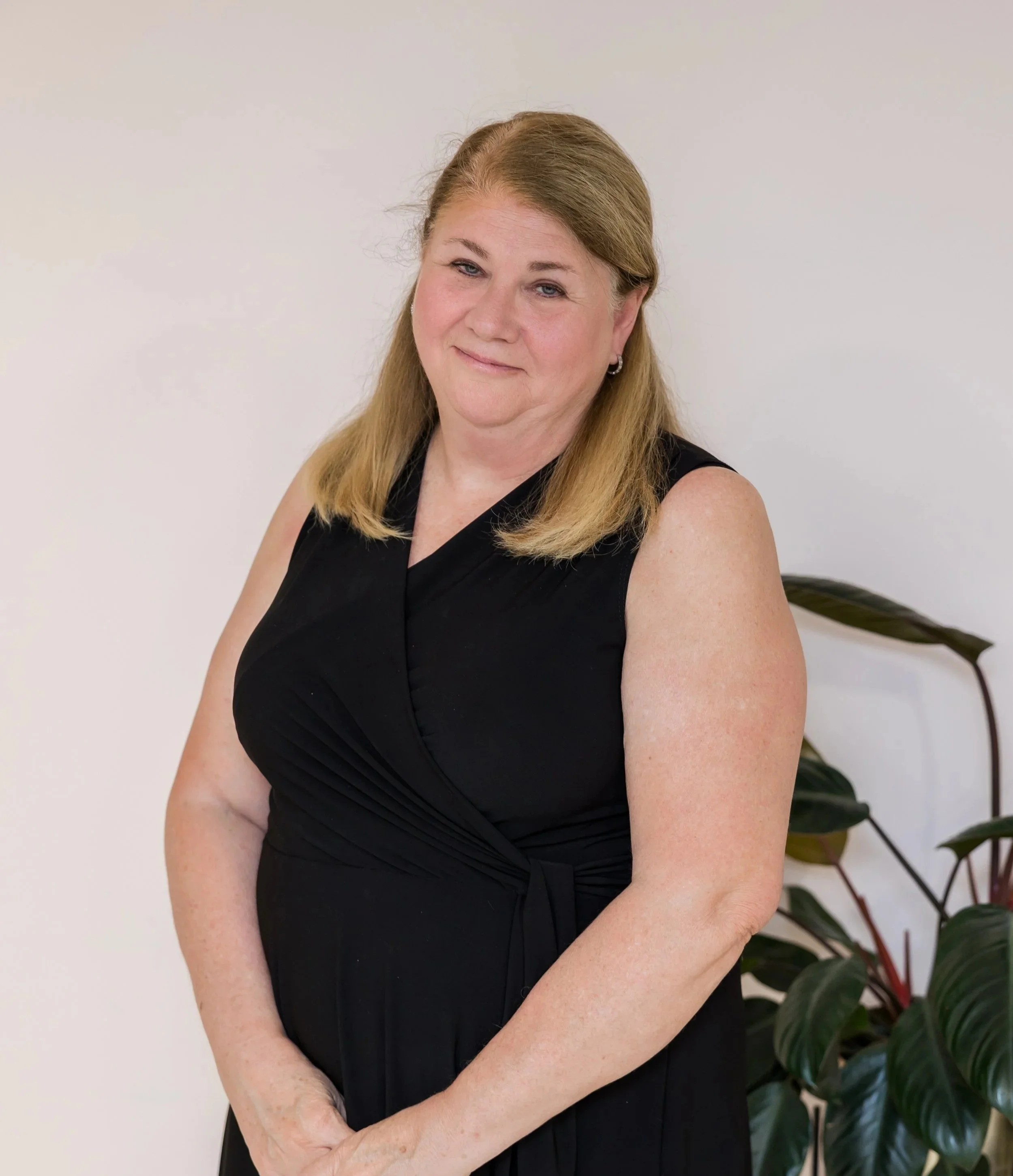 A middle-aged woman with shoulder-length blonde hair wearing a black sleeveless dress, standing against a plain white wall with a large potted plant to her right.