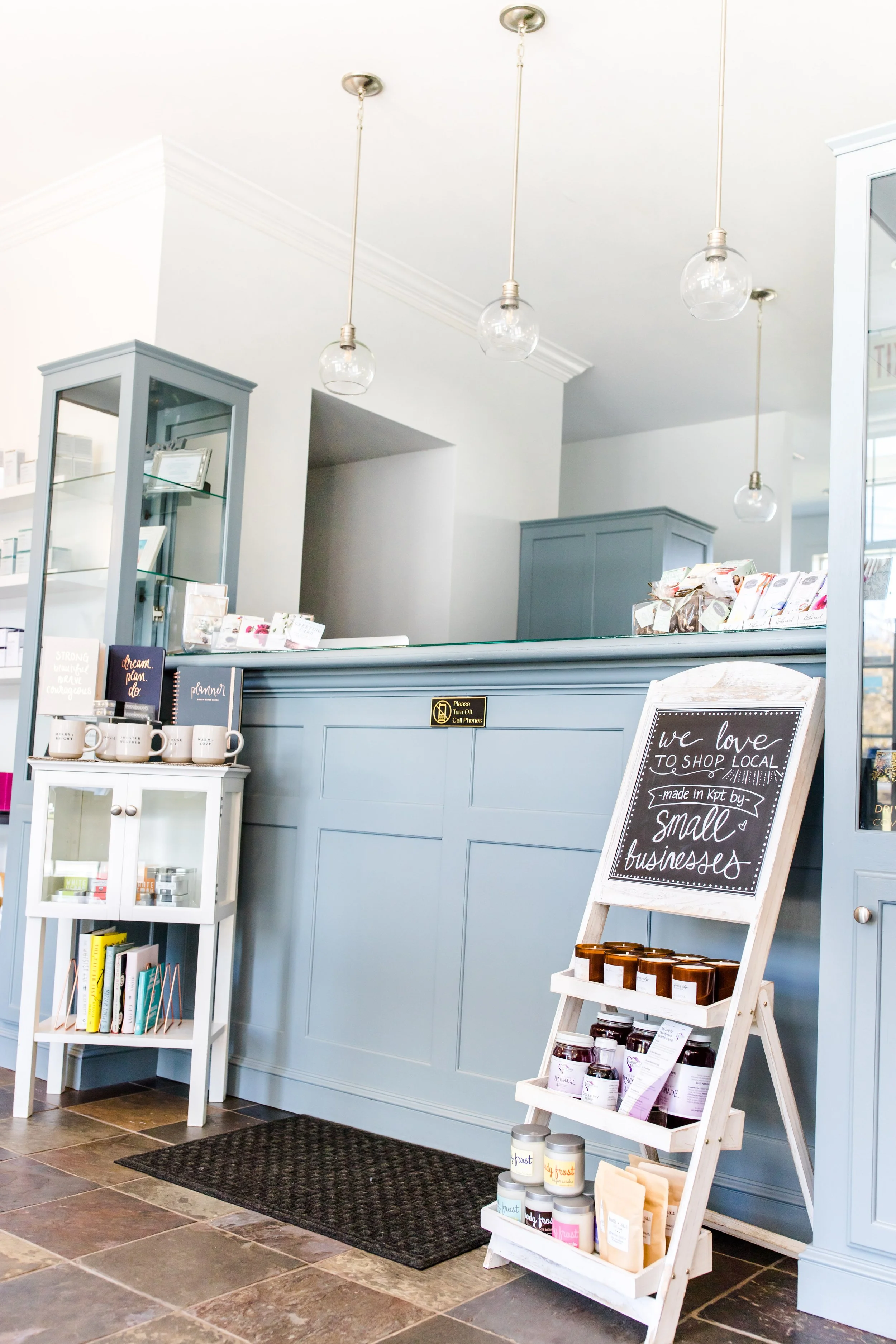 Interior of a small retail shop with a wearing counter. There are small shelves with products, a chalkboard sign promoting local small businesses, and pendant lights hanging from the ceiling.