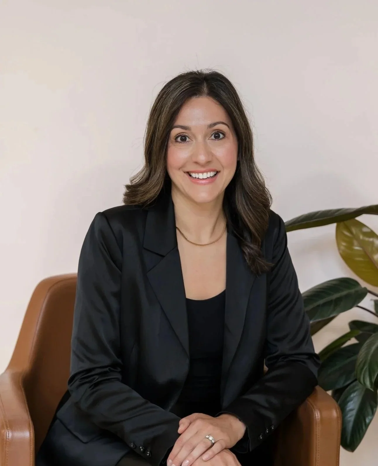 A woman with shoulder-length brown hair, wearing a black blazer and black top, sitting on a tan leather chair, smiling at the camera.