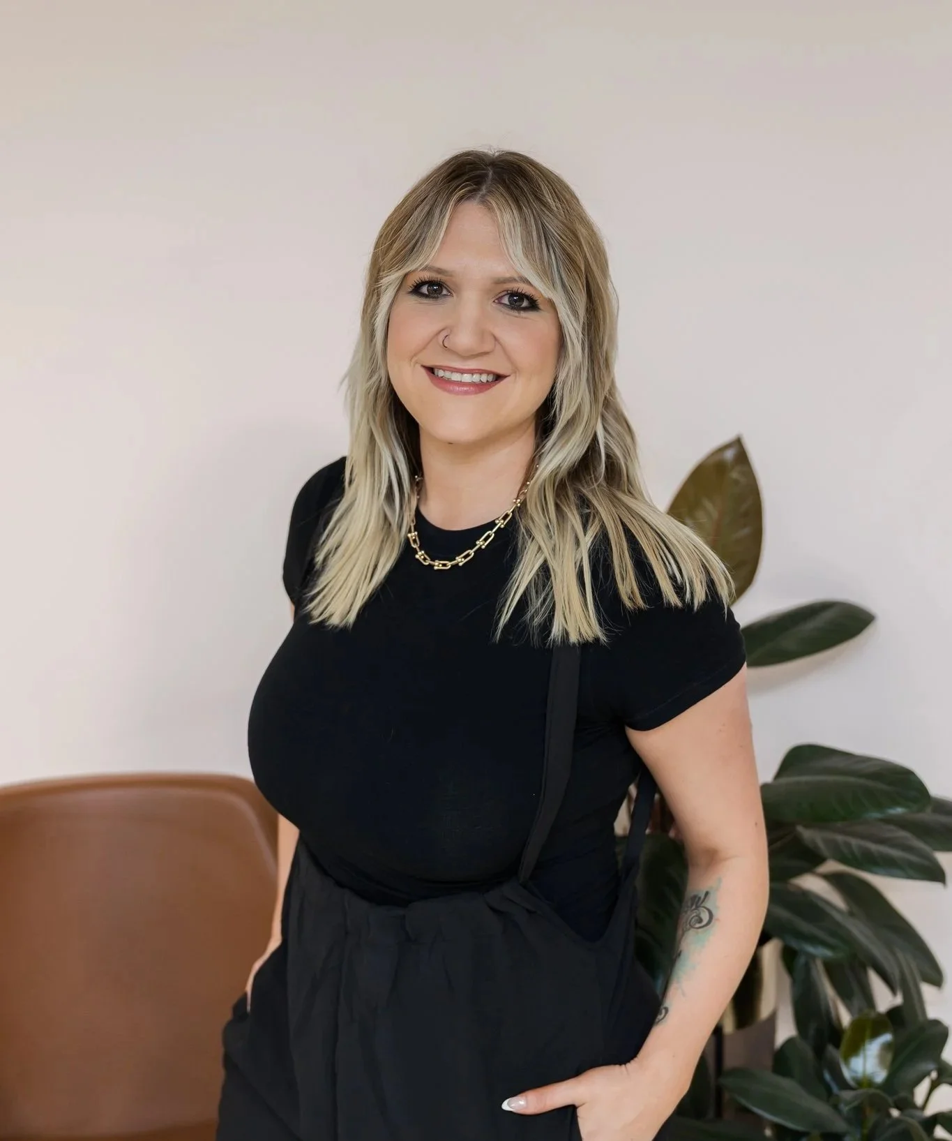 A smiling woman with blonde hair wearing a black shirt and gold chain necklace, standing in front of a white wall and green plant.