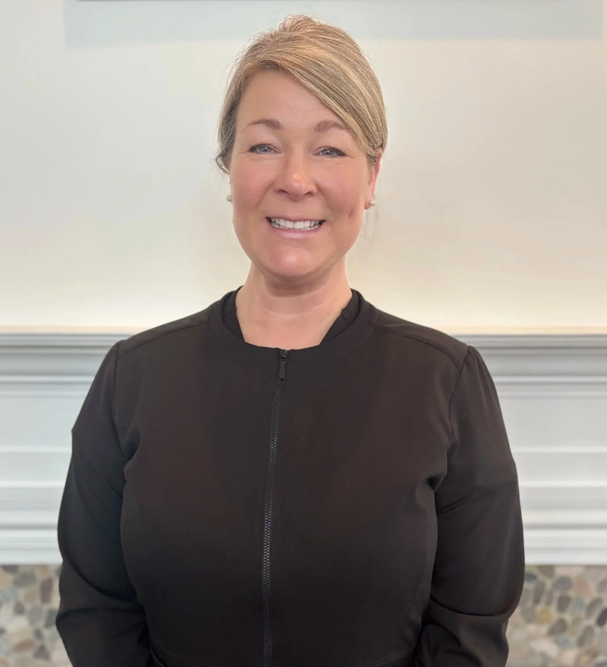 A woman with short blonde hair smiling, wearing a black zip-up jacket, standing in front of a white wall with a decorative molding.