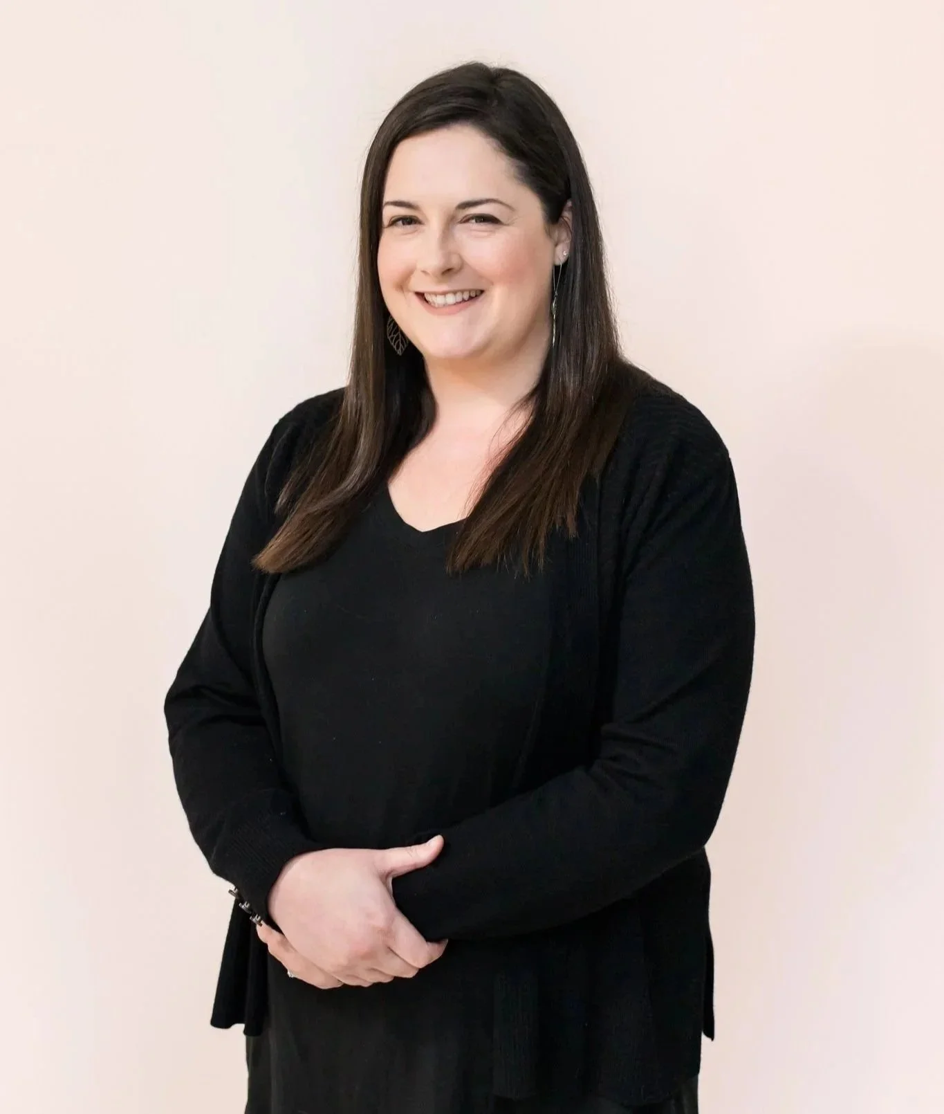 A woman with long dark hair, smiling, wearing a black top and black cardigan, standing against a plain light-colored background.