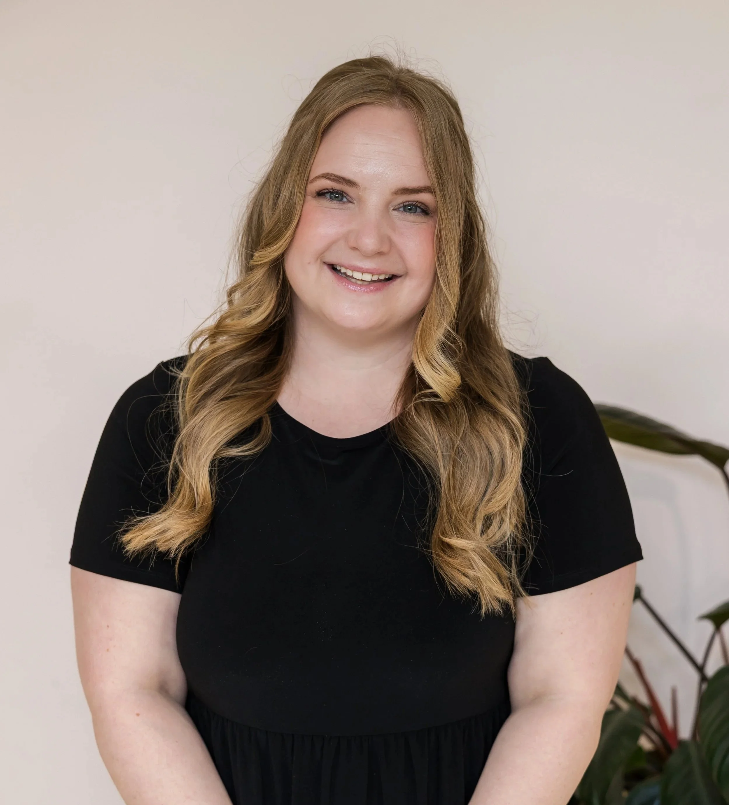 A woman with long wavy blonde hair smiling, wearing a black top, sitting in front of a plain off-white wall with a plant in the background.