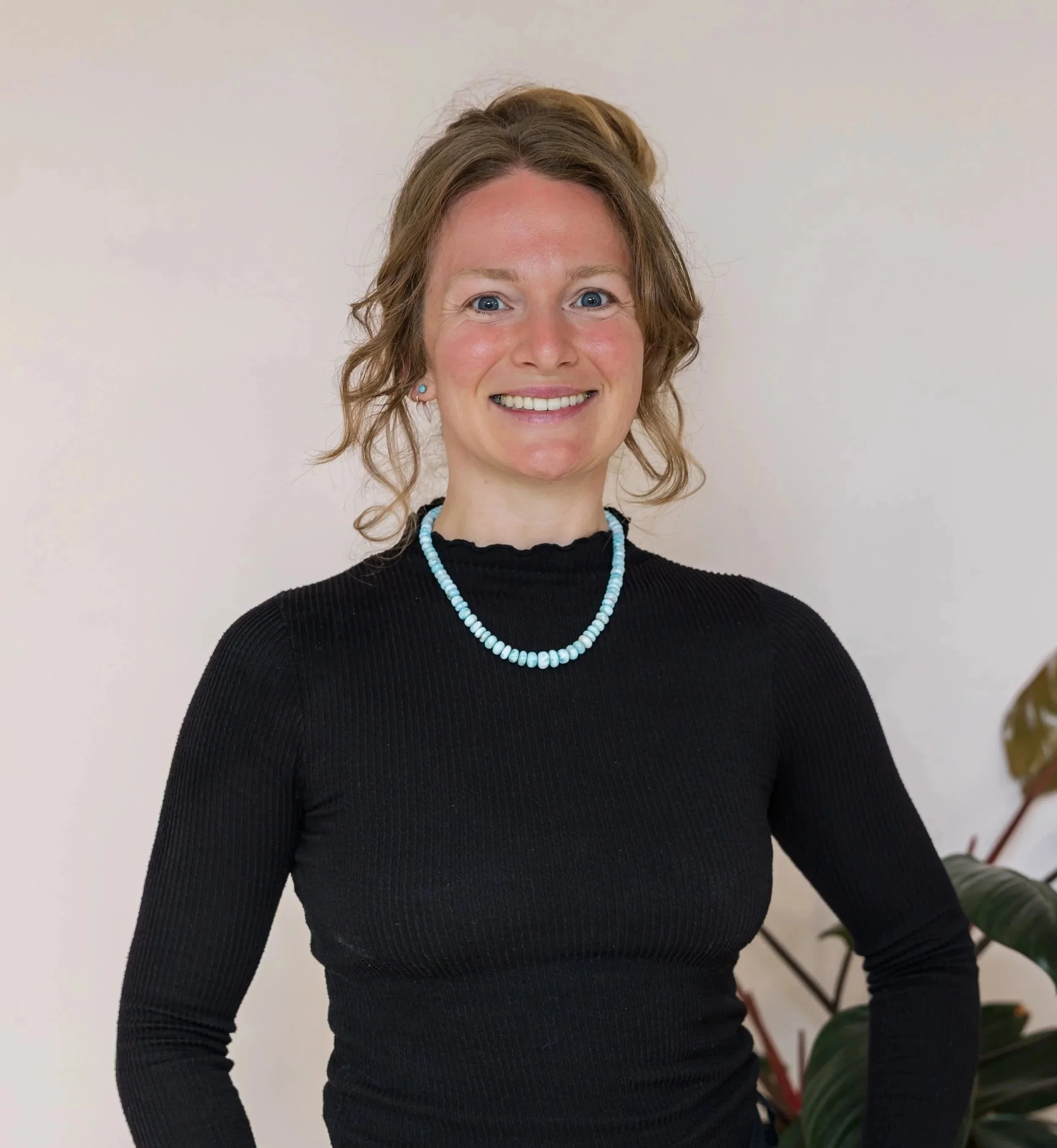 A woman with light skin and curly, shoulder-length hair tied in a styled bun, smiling and posing against a plain light-colored wall, wearing a black long-sleeve top and a light blue beaded necklace.