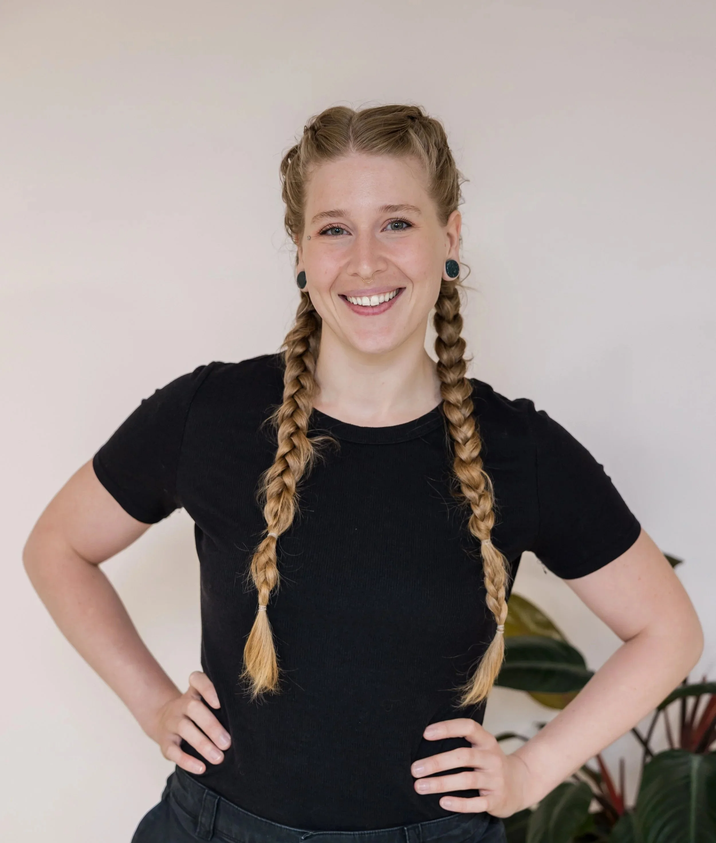 A young woman with braided hair, smiling, wearing a black t-shirt and standing with hands on hips in front of a plain light-colored background.