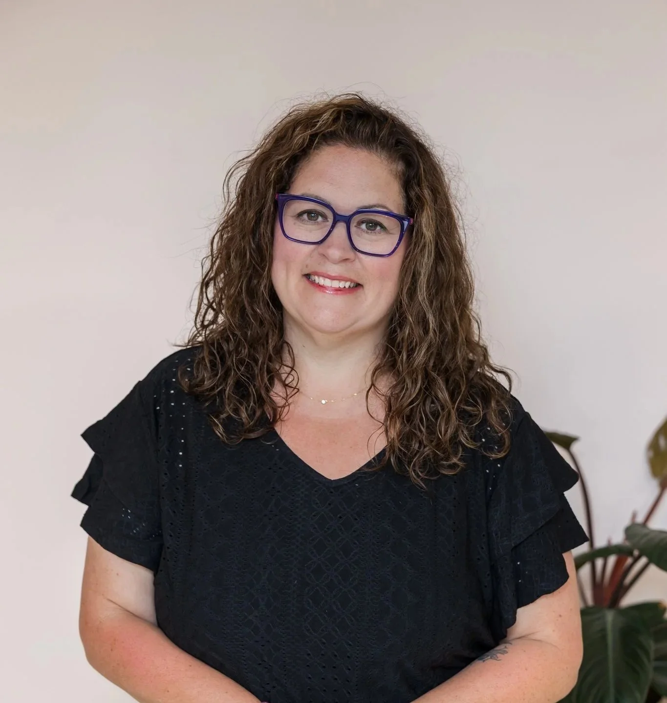 A woman with curly brown hair wearing purple glasses and a black dress, smiling, standing in front of a light neutral background.