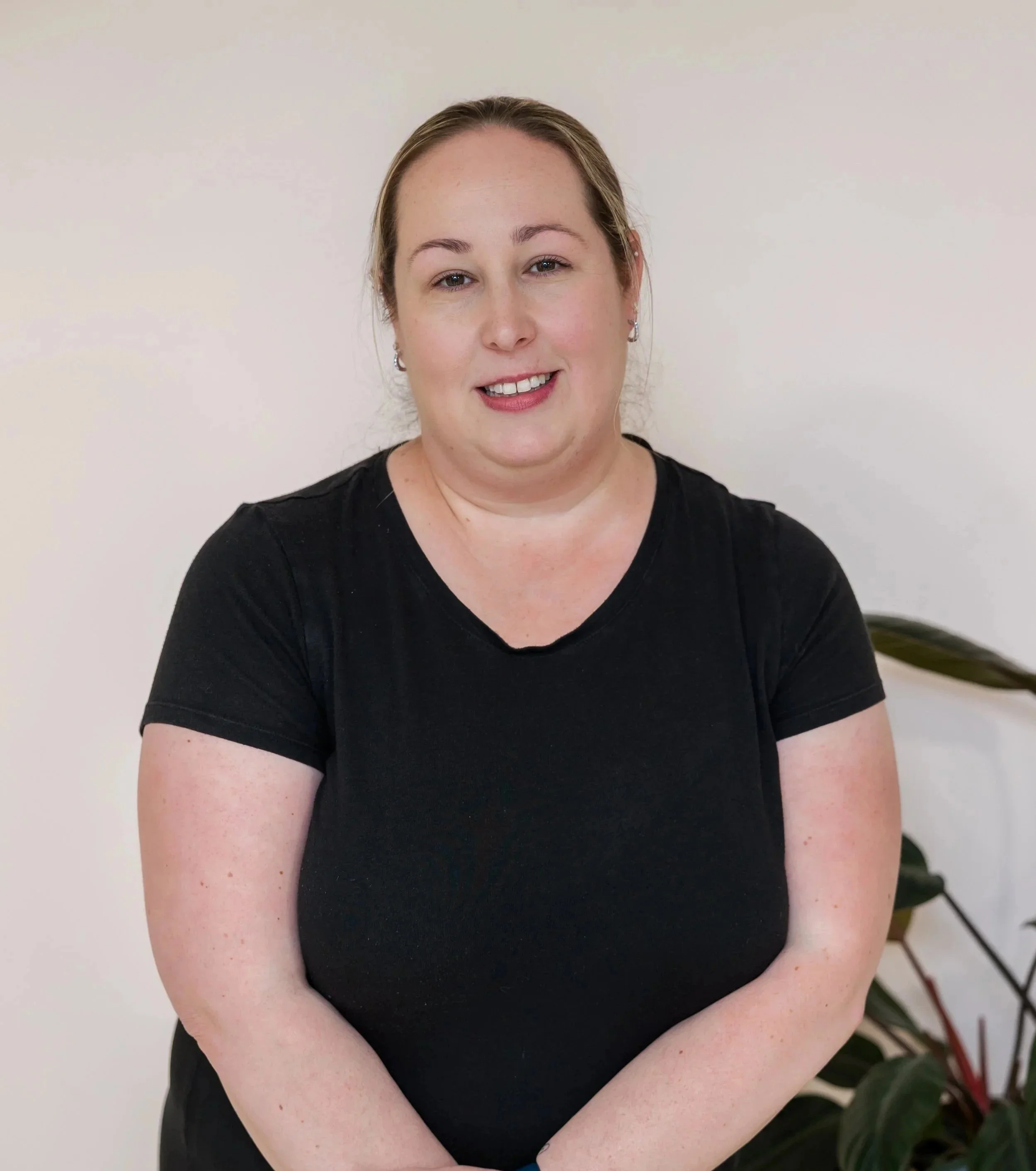 A woman wearing a black t-shirt standing against a plain white wall, smiling gently, with a plant visible to her right.