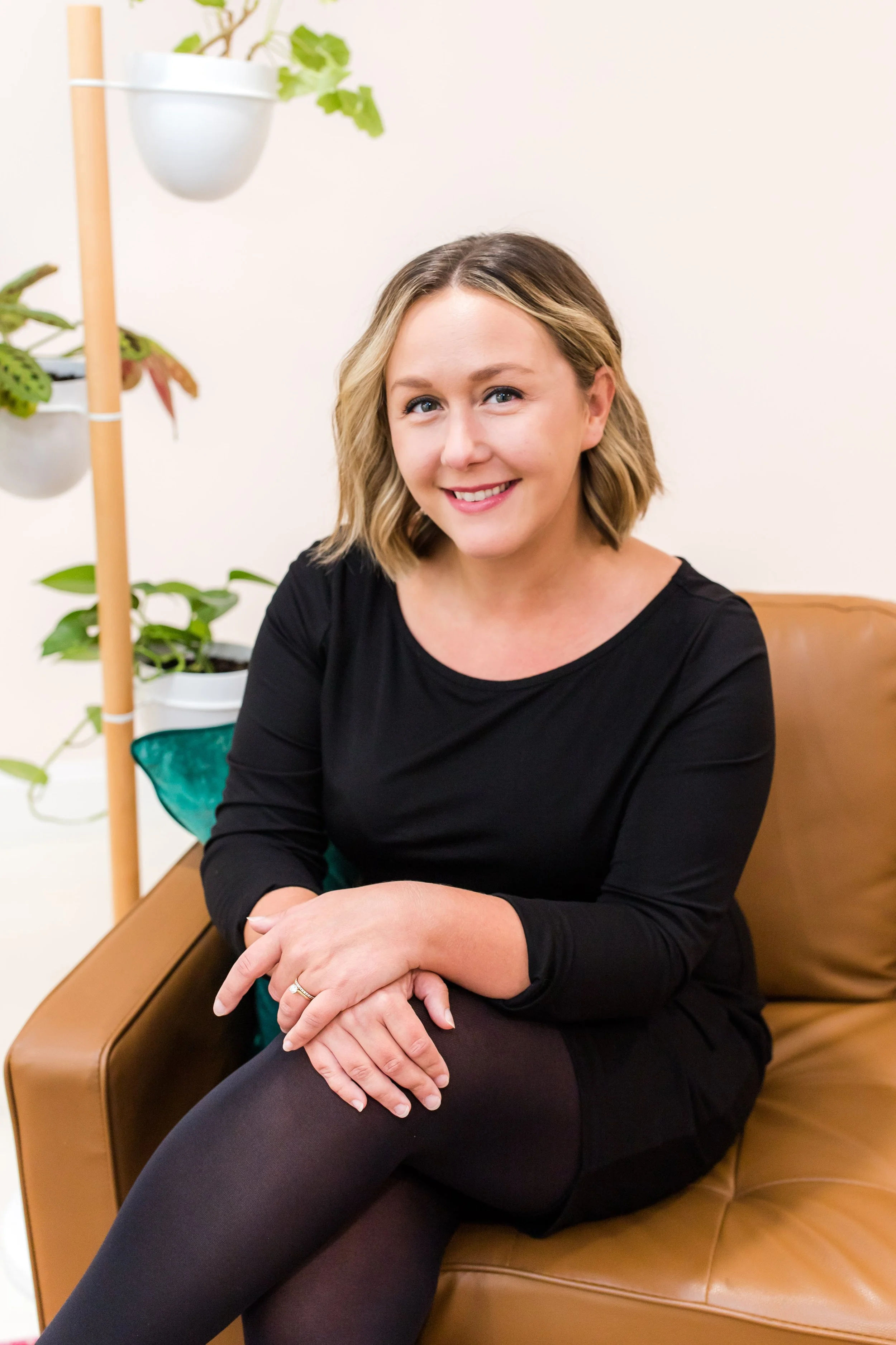 A smiling woman with shoulder-length blonde hair, wearing a black top and black tights, sitting on a tan leather chair in an indoor setting with potted plants in the background.