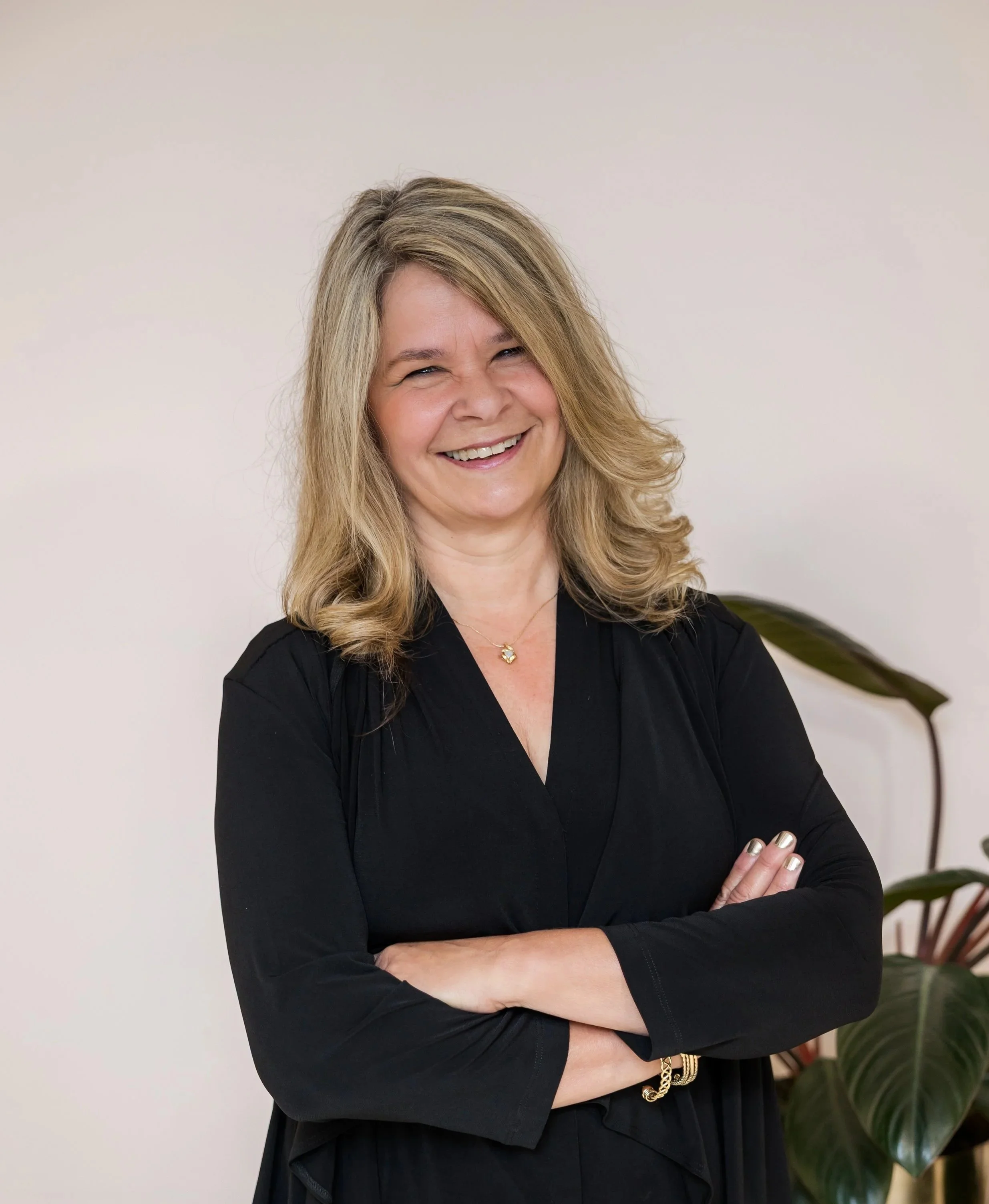 A woman with blonde hair smiling, wearing a black top, standing with her arms crossed in front of a white wall and a potted plant.