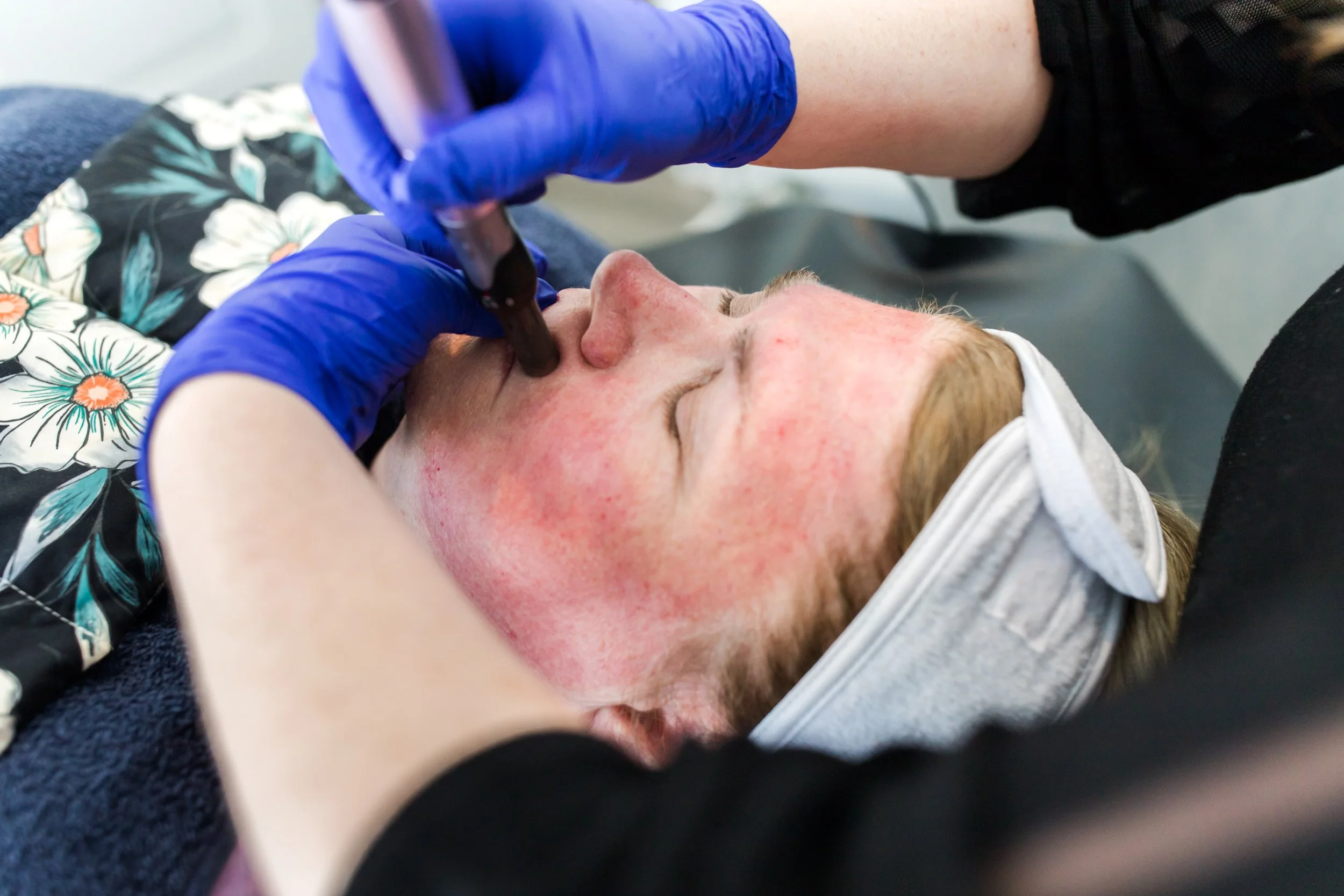 A person wearing a floral shirt and a face mask receiving a tattoo on their face from an artist with blue gloves.