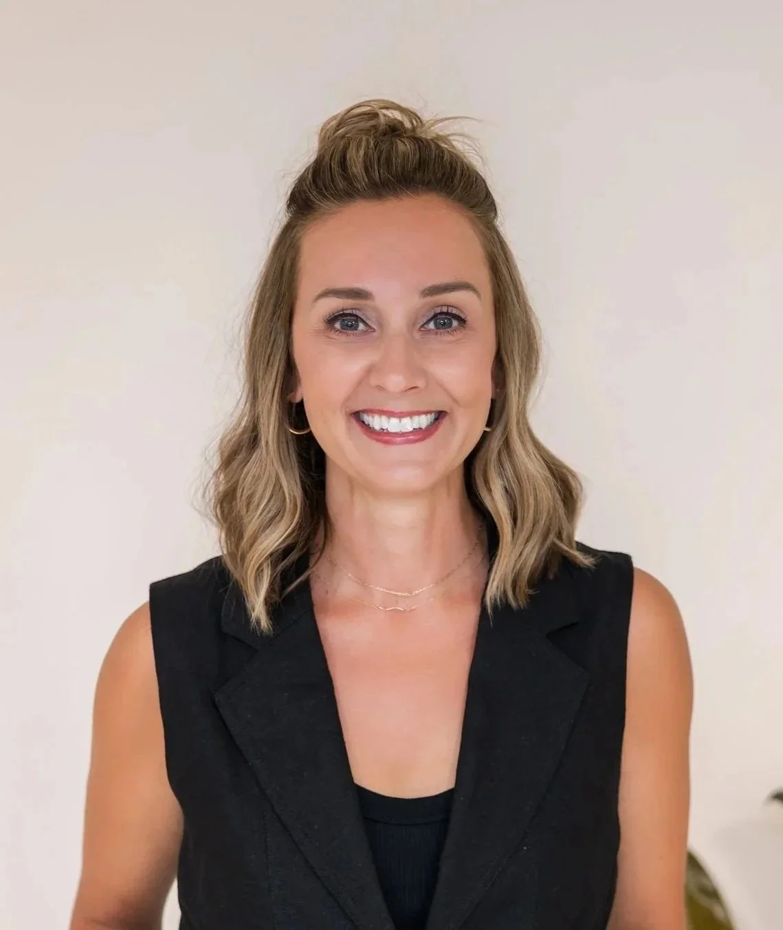A woman with light brown, shoulder-length wavy hair styled with a top knot, smiling, wearing a black sleeveless blazer and a tiny gold necklace, standing against a plain off-white wall.