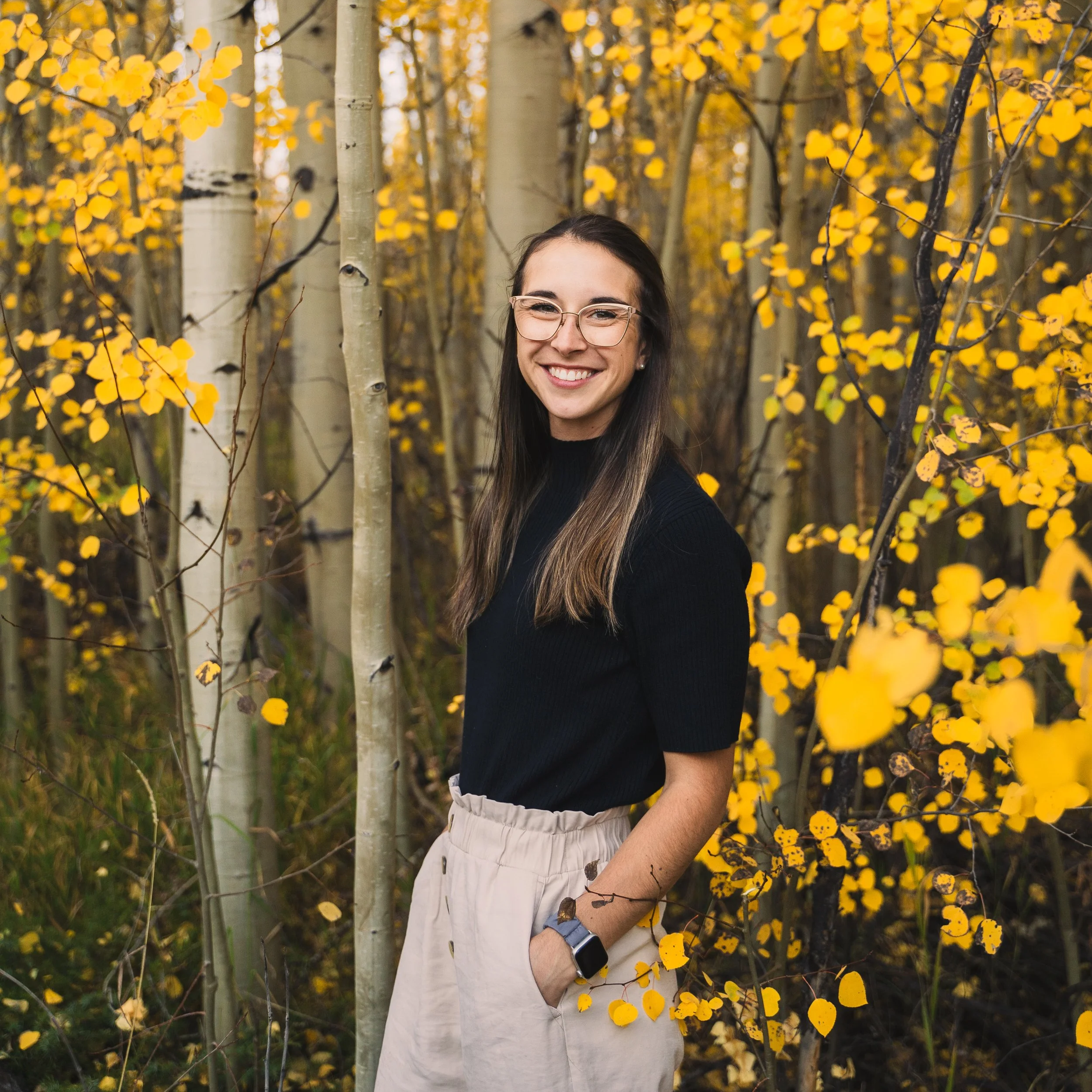 A young woman with long dark hair, glasses, and a black shirt is smiling in a wooded area with yellow autumn leaves on trees and branches.