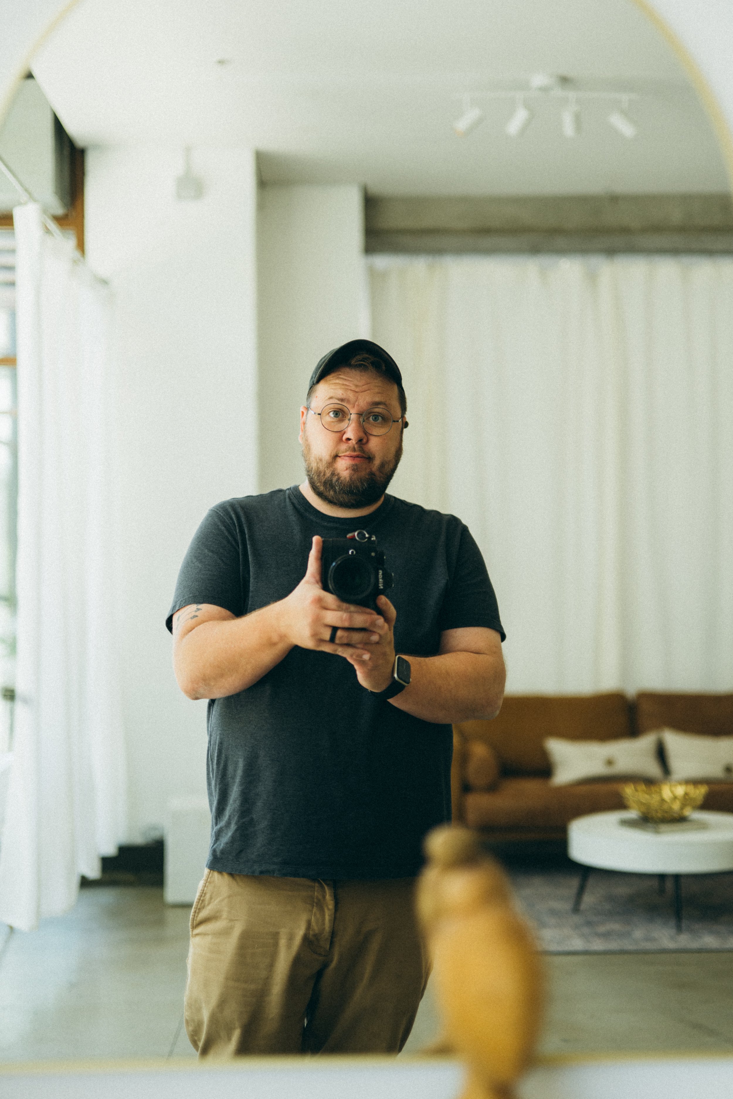 A man taking a selfie in front of a mirror in a living room with a beige couch and white curtains.