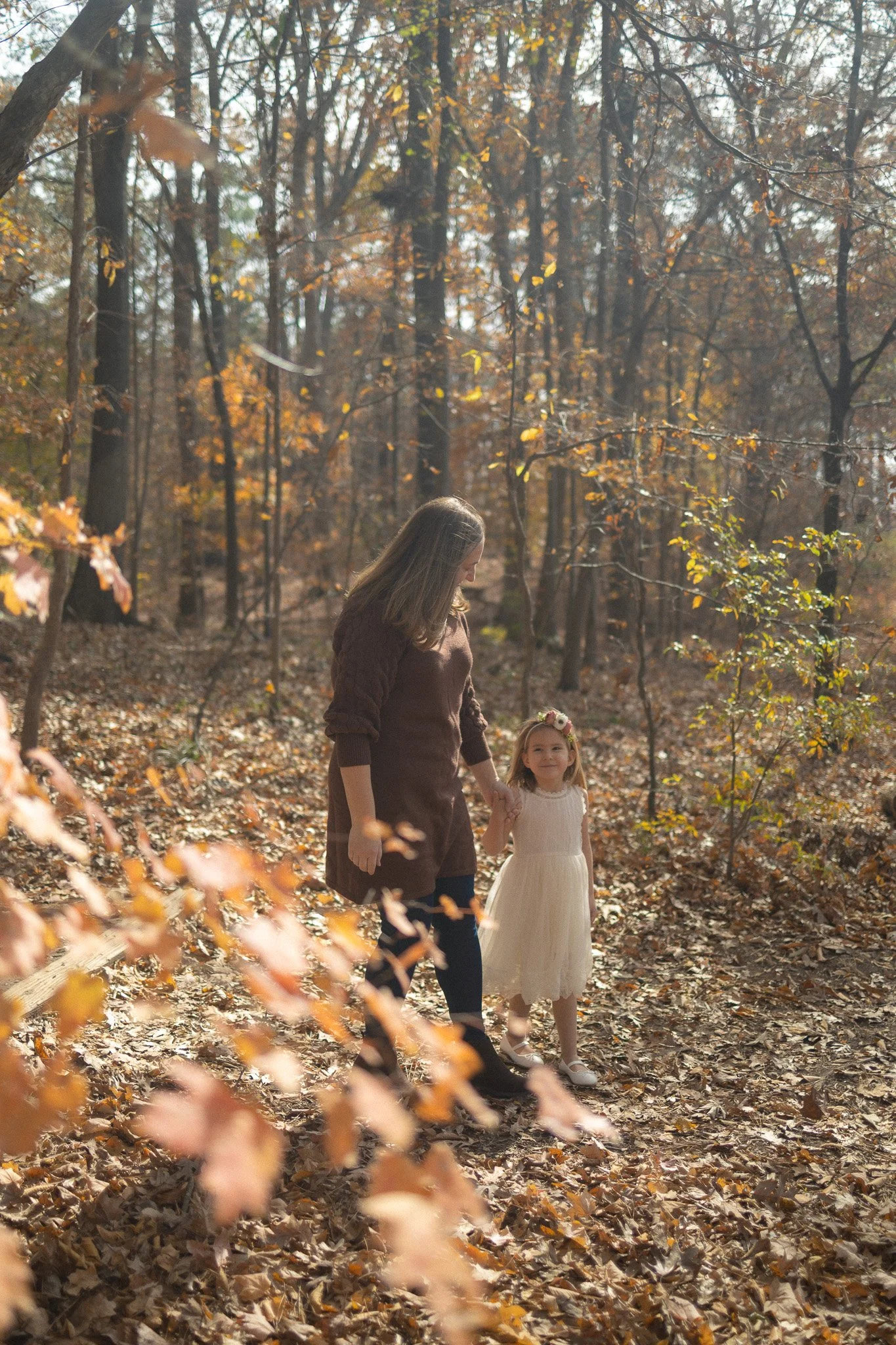 Mother and young daughter holding hands walking away through a sunlit autumn forest in Morrisville, NC.