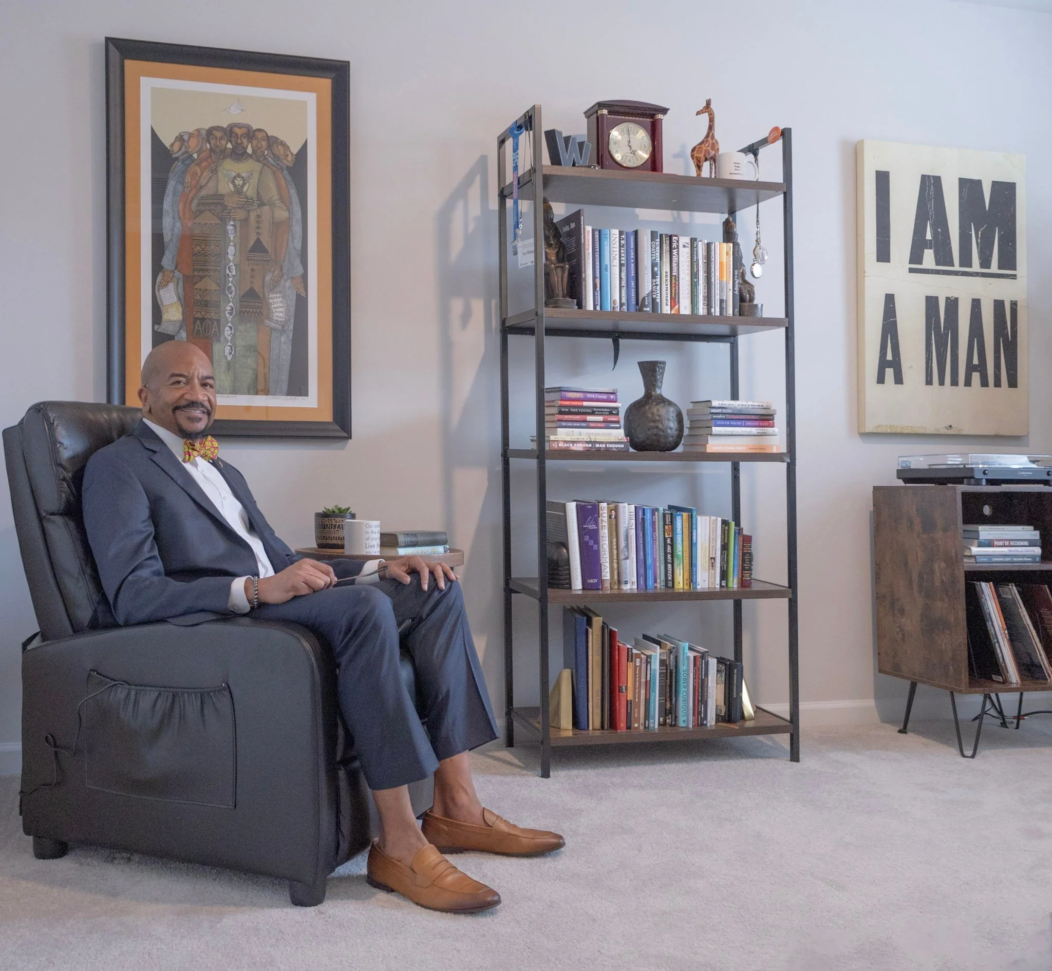 Environmental portrait of a Black male leader in a suit and bowtie, seated in a leather recliner next to a bookshelf and art in a Durham home office.