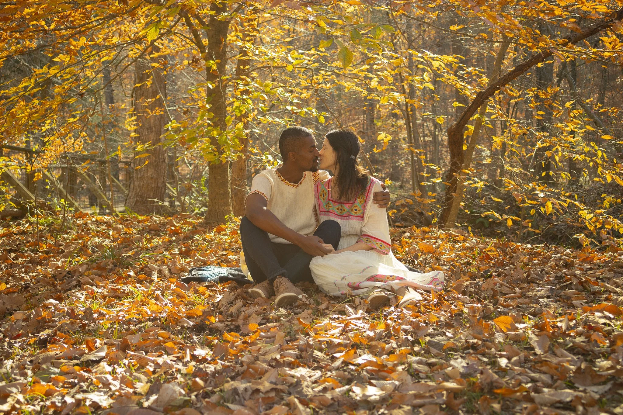 Candid lifestyle shot of an interracial couple sitting on autumn leaves in a sunlit forest, sharing an intimate moment.