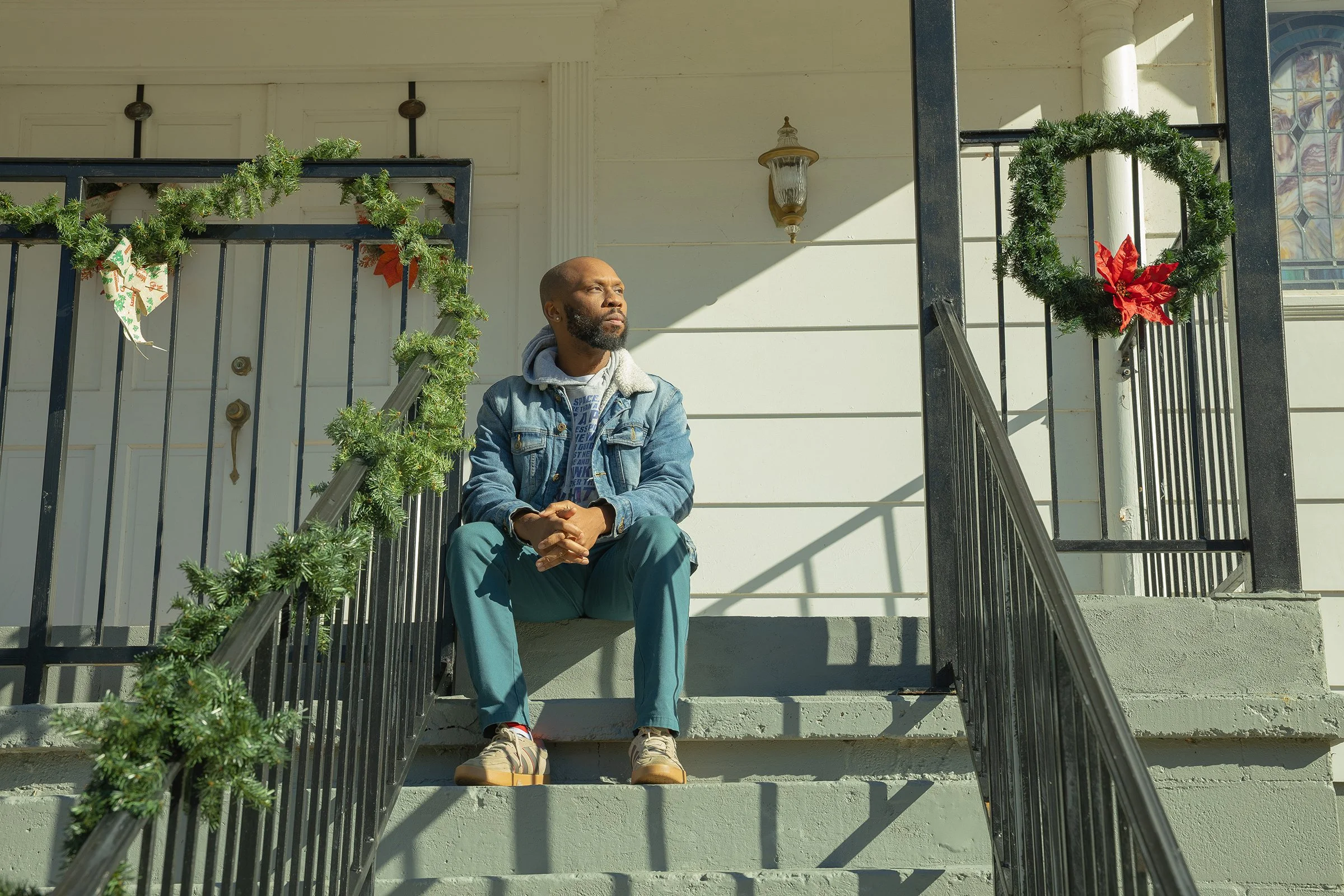 Editorial portrait of a Black man in a denim jacket sitting on the concrete steps of a white church with holiday wreaths, looking away from the camera.