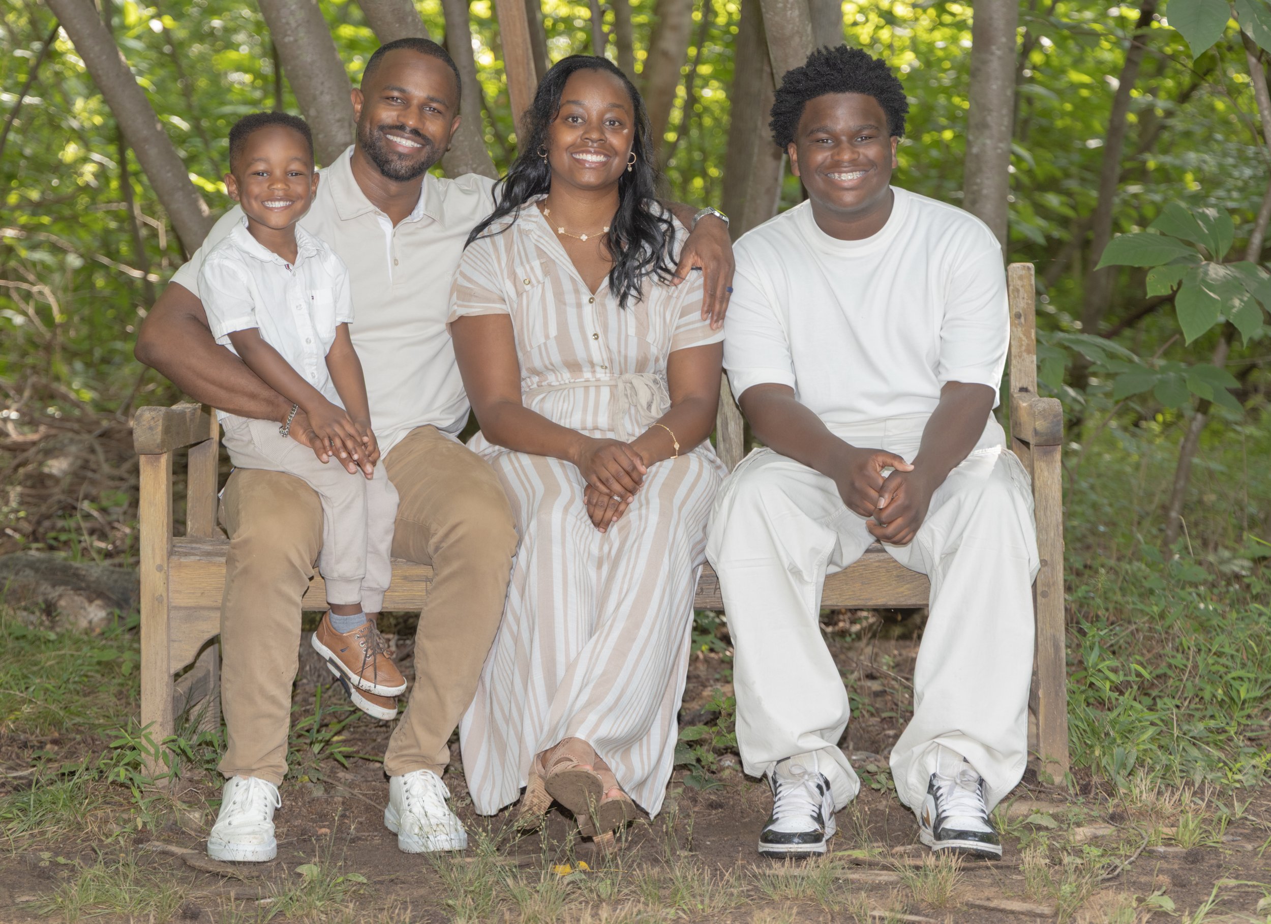 Durham NC outdoor family photographer. A smiling family of four sitting together on a wooden bench in a lush, green forest setting, wearing coordinated neutral tones. Environmental family portrait by Kwaku Osei Studios.