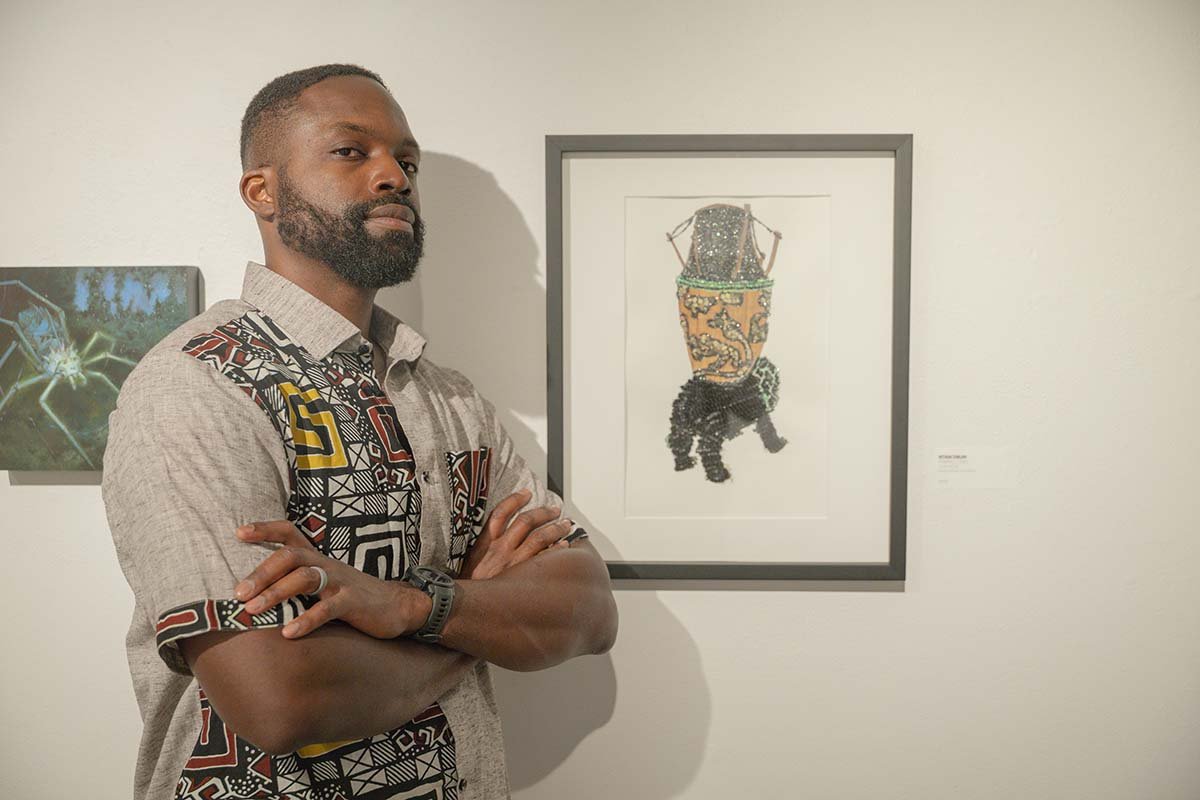 Portrait of artist Kwaku standing with his arms crossed next to his framed artwork in a gallery setting. He is wearing a gray shirt with African geometric patterns. The artwork behind him features a highly textured, beaded drum on an animal figure.