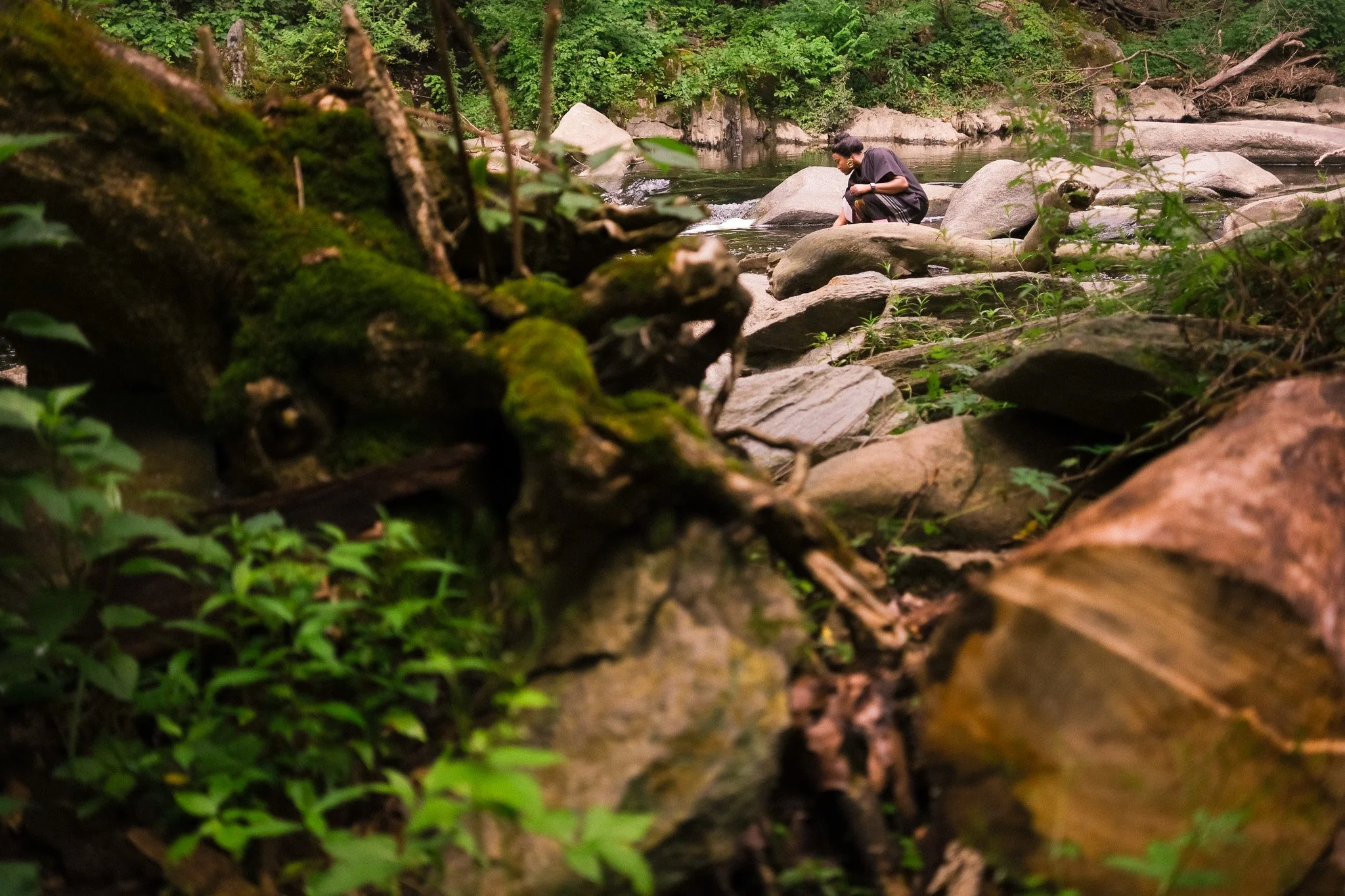 Environmental portrait photographer capturing a subject by a rocky riverbank in a lush, green forest.