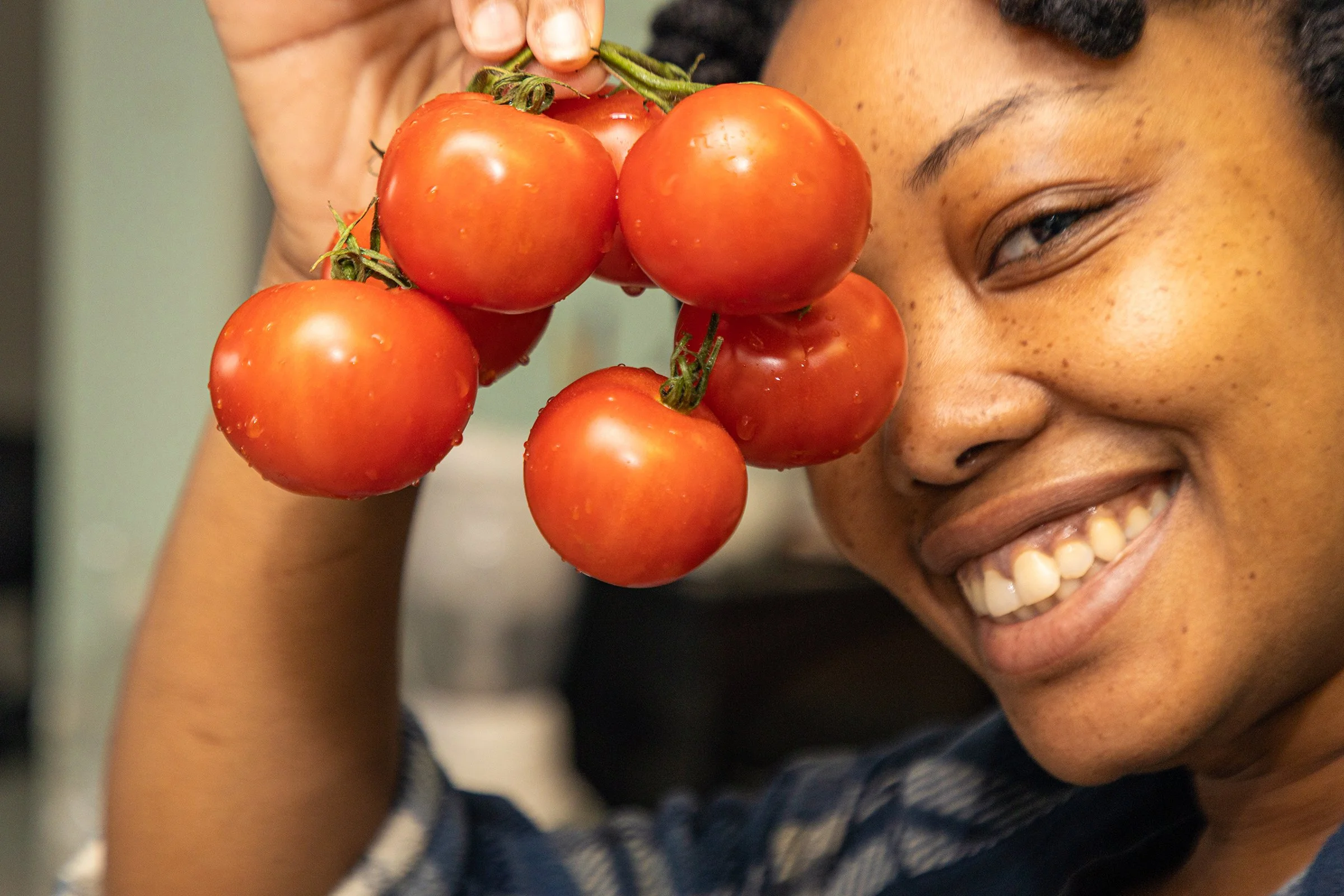 A smiling Black woman holds fresh vine tomatoes close to her face in a warm kitchen. This environmental portrait by Ade Carrena photography highlights joyful Black portraiture.